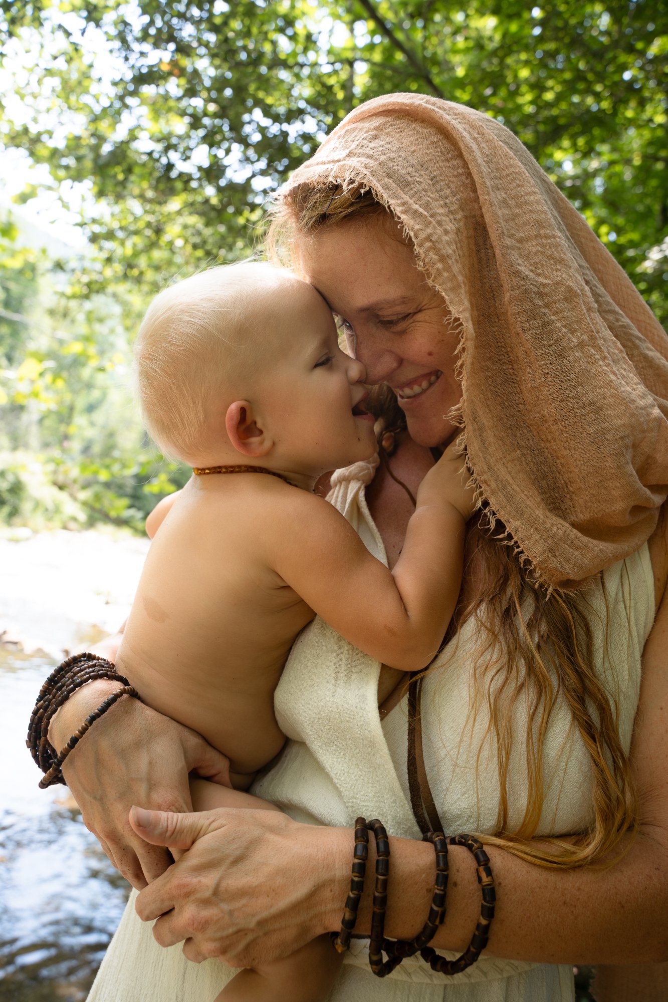A woman holding a young child close, both smiling and touching foreheads in an outdoor forest setting.