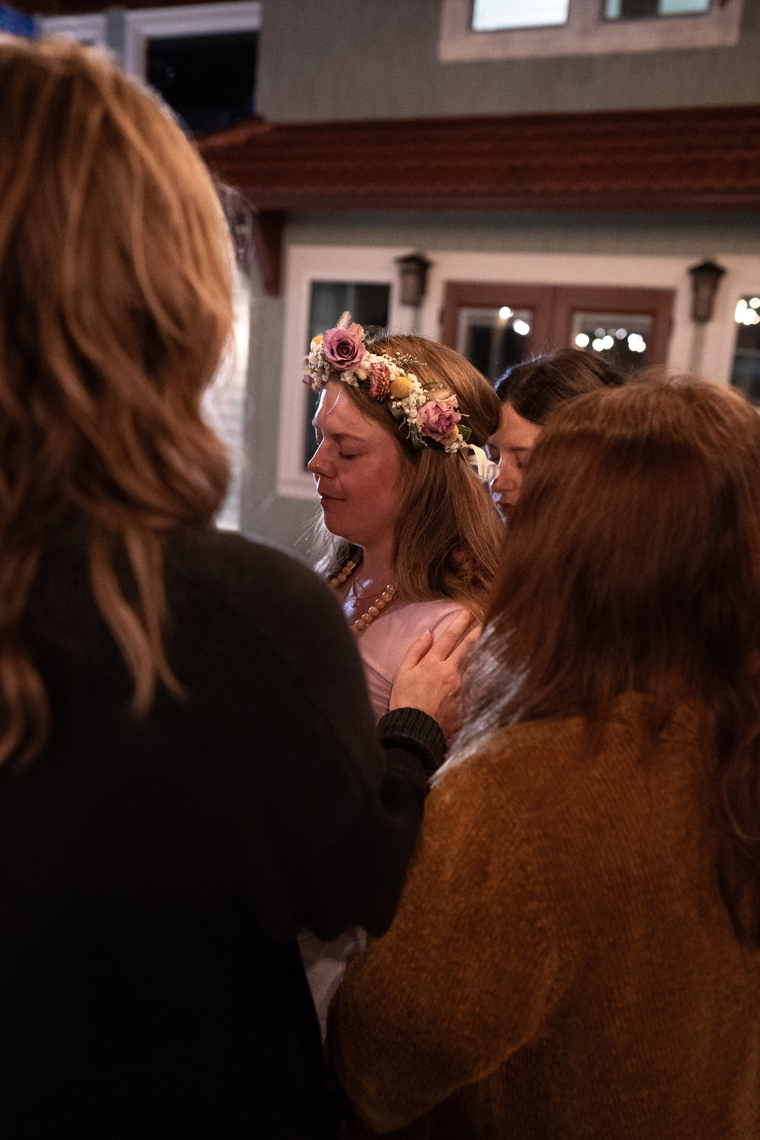 A woman with a floral crown praying with her eyes closed surrounded by friends during a heartfelt moment.