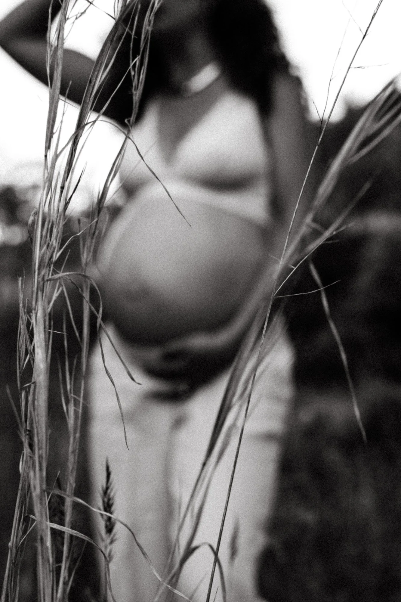 A pregnant woman standing outside among tall grass, looking down, blurred in the background, black and white photo.