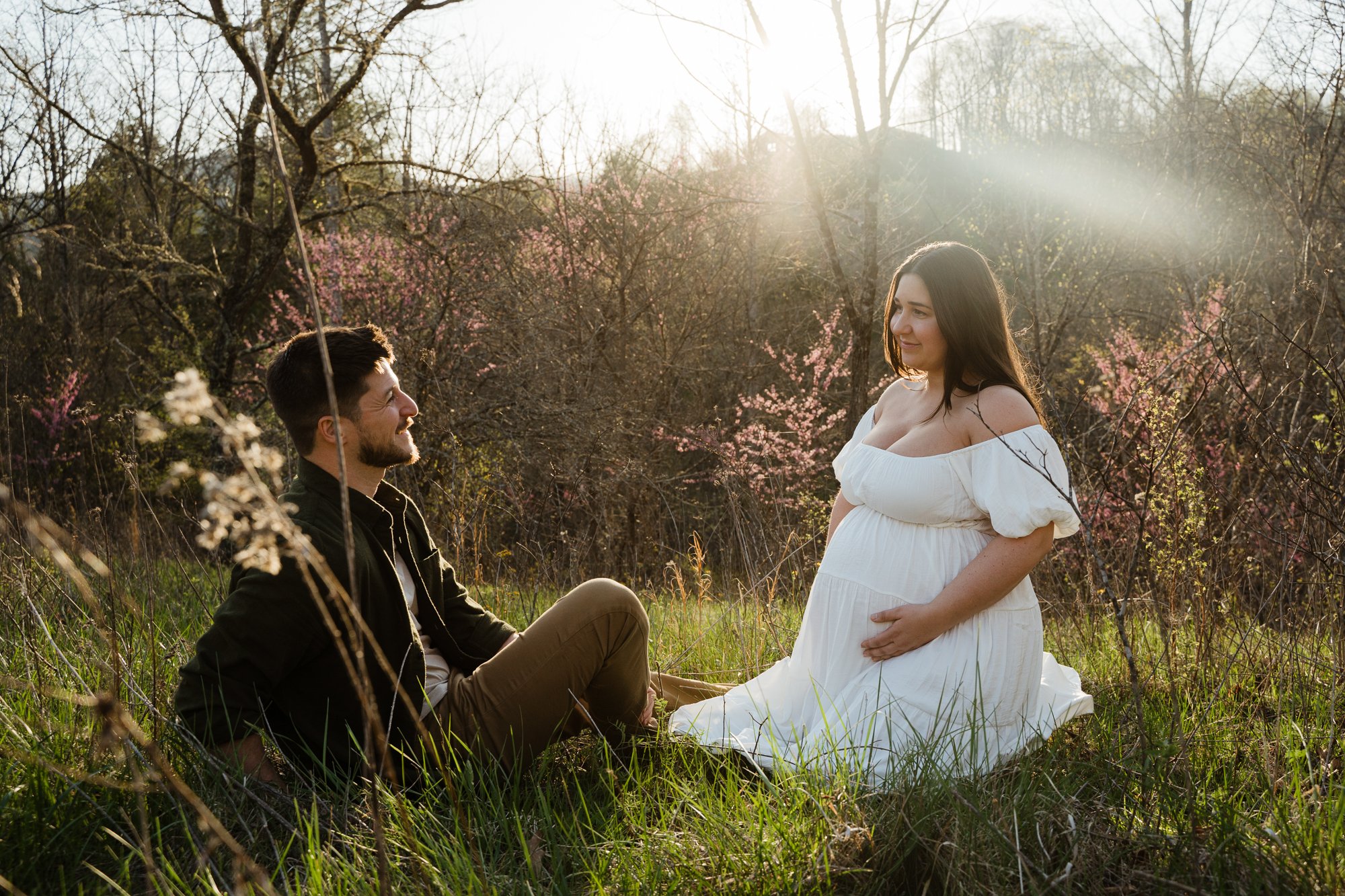 A man sitting on the grass looking at a pregnant woman in a white dress, in a field with blooming trees and sunlight shining through.