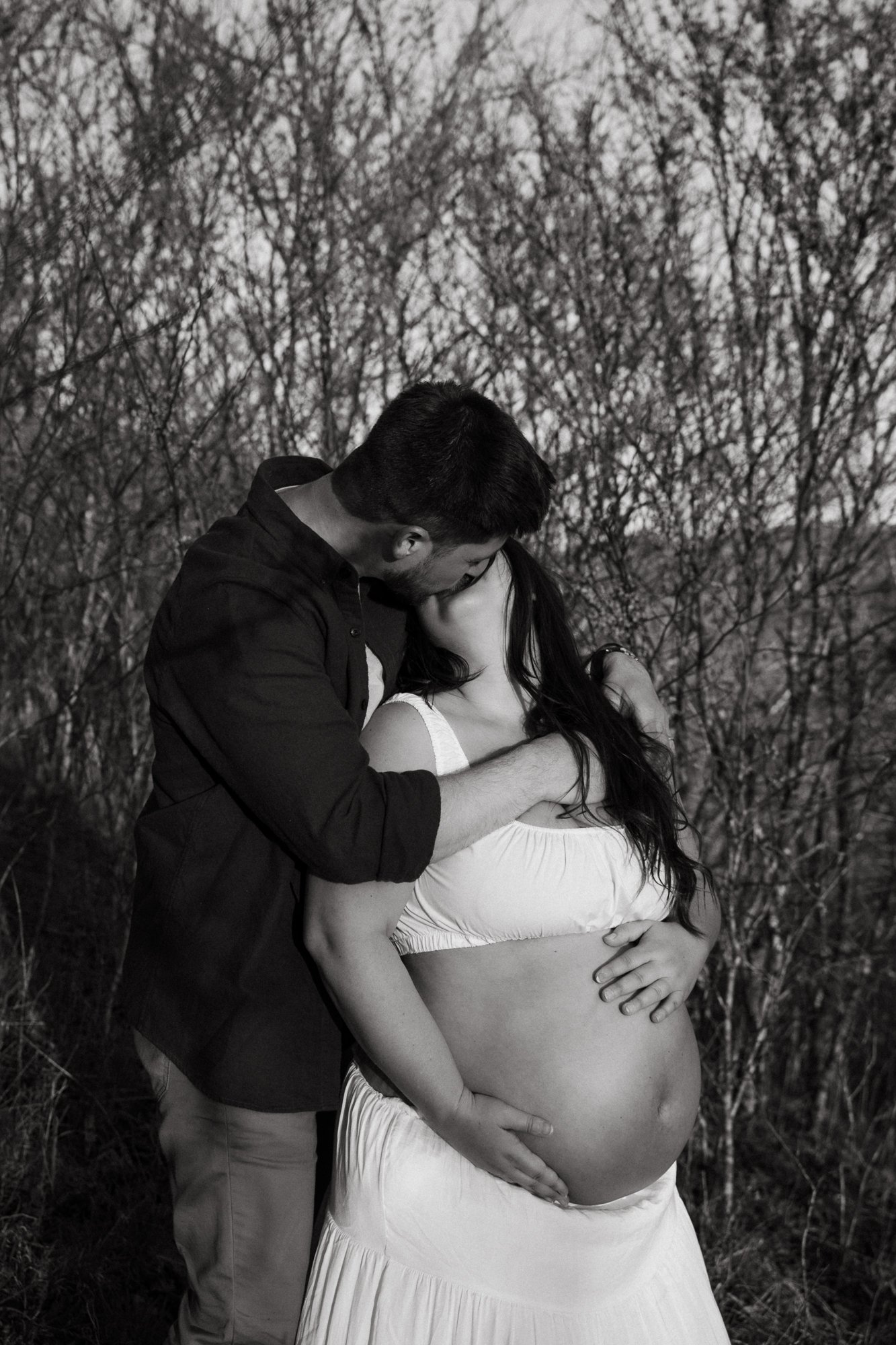 A black and white photo of a pregnant woman and a man kissing outdoors, with the man holding the woman's face and her hand resting on her belly, surrounded by bare trees.