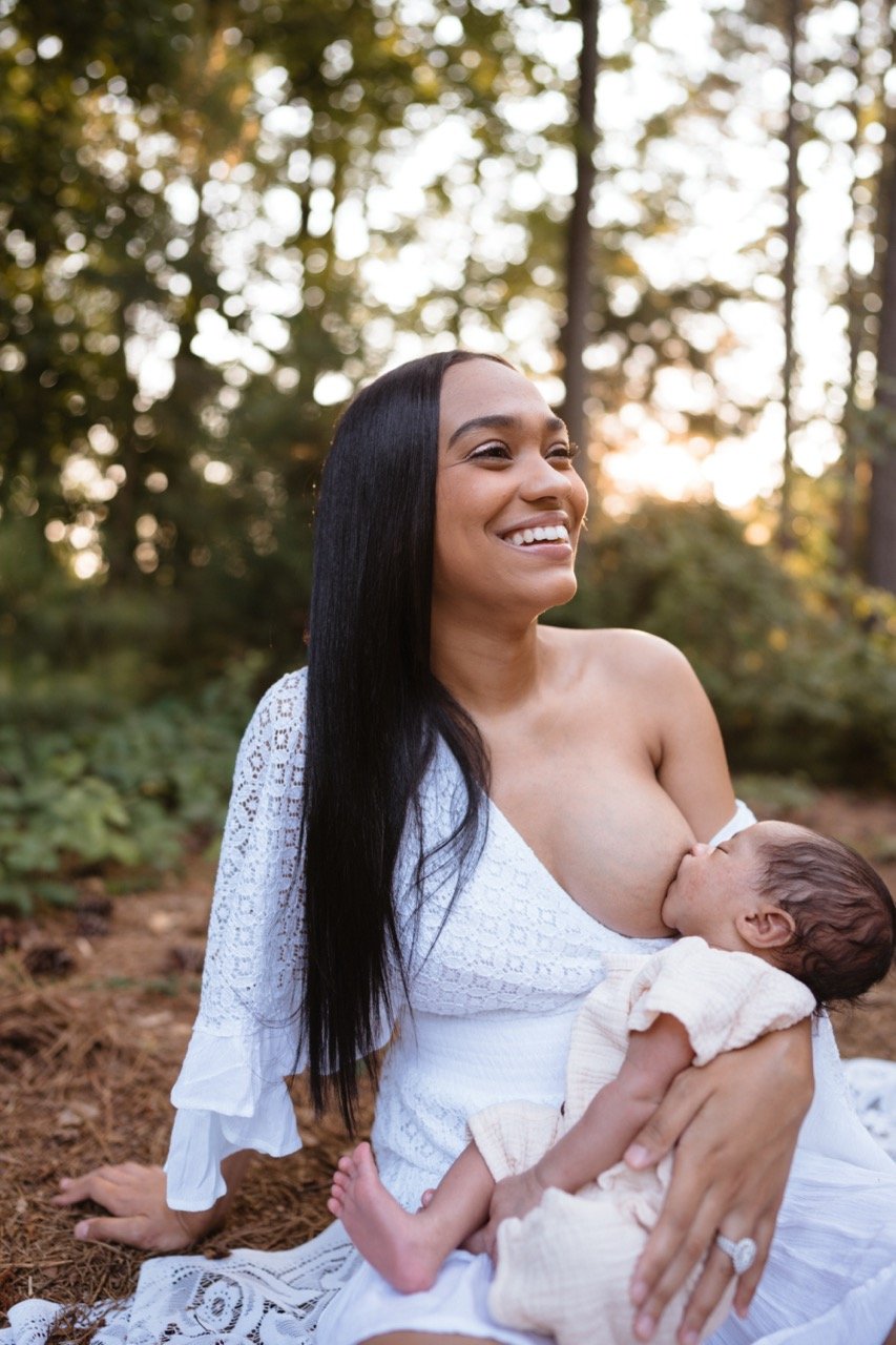 A woman with long dark hair, wearing a white lace dress, is breastfeeding a baby outdoors in a forest setting at sunset, smiling happily.