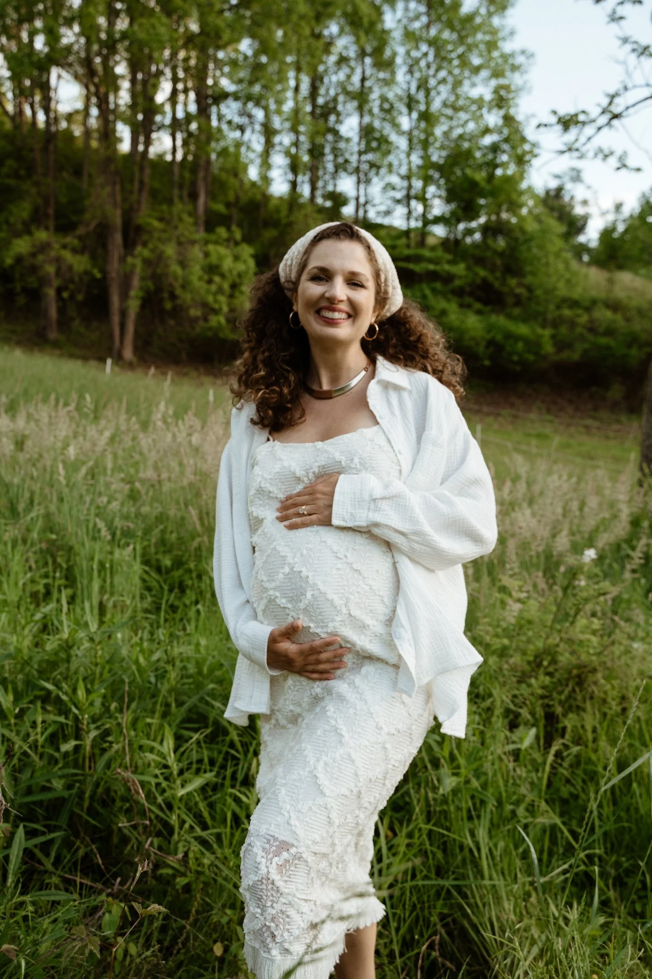 A pregnant woman with curly hair smiling while standing in a field of tall green grass surrounded by trees, dressed in a white lace dress, white shirt, and accessorized with hoop earrings, a necklace, and a headband.