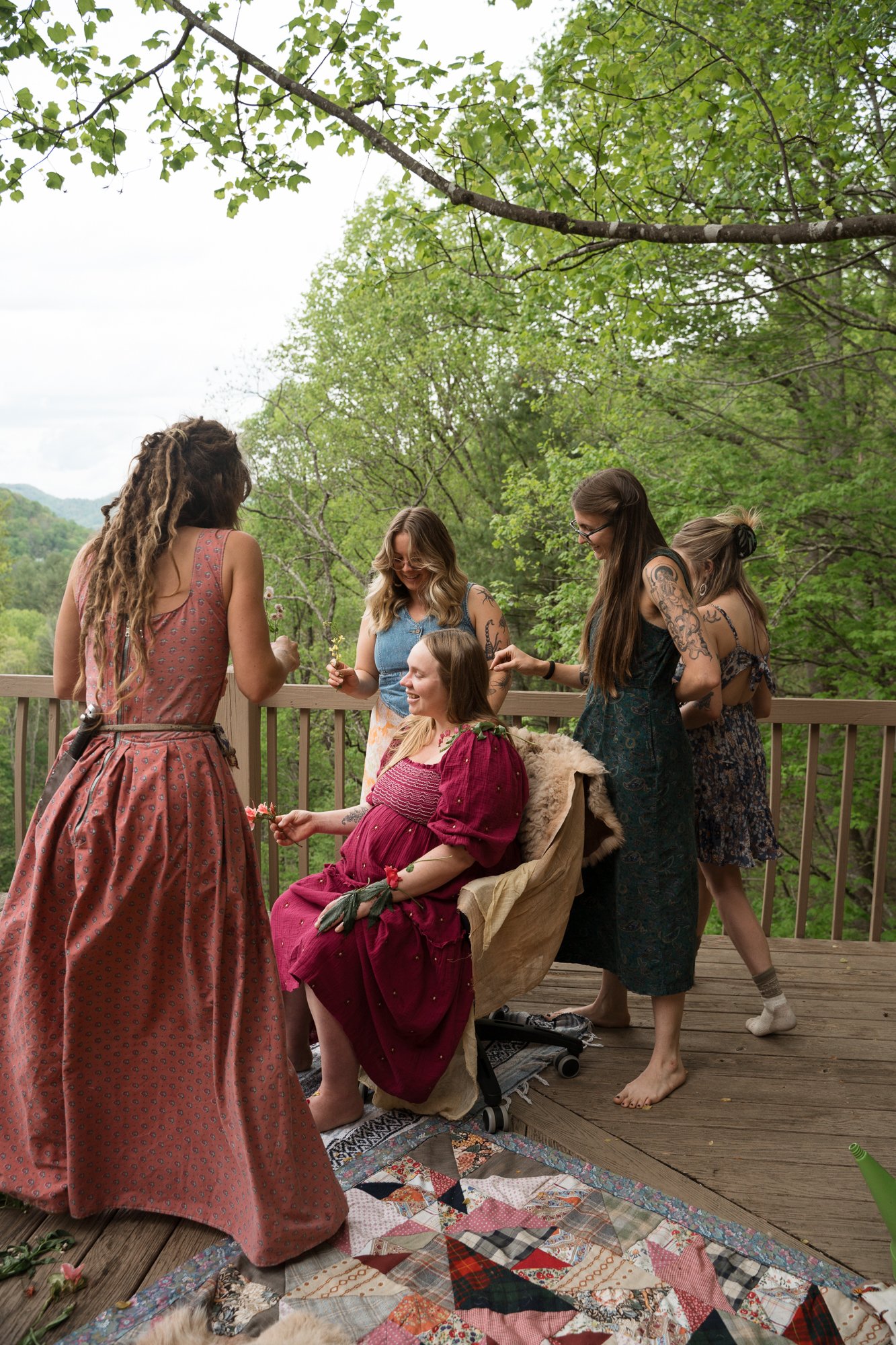 Group of women celebrating a woman in a red dress on a wooden deck surrounded by green trees.