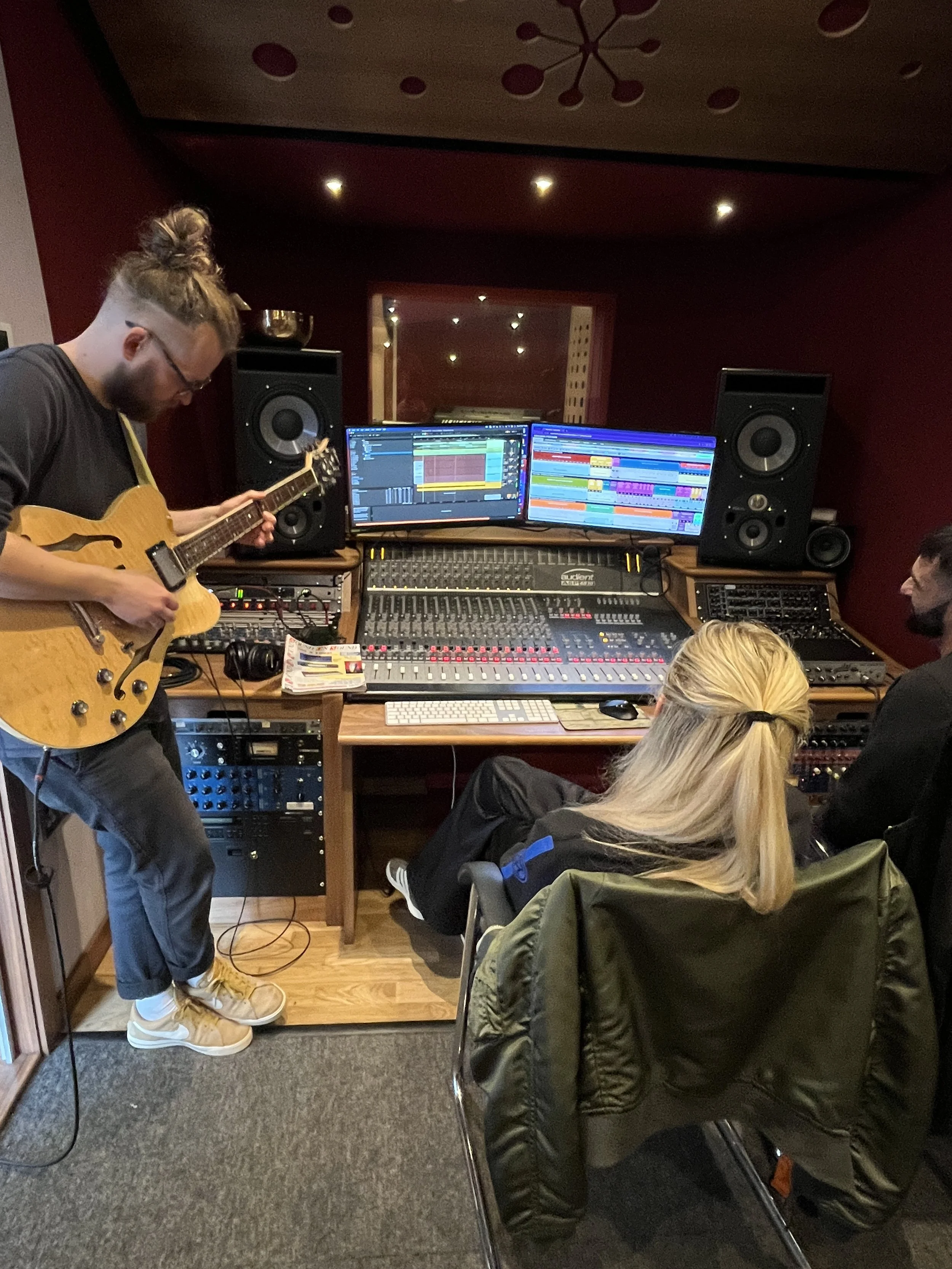 A person with glasses and a bun hairstyle playing an electric guitar in a recording studio. There are two computer screens with music production software, large speakers, a mixing console, and other recording equipment. Several people are sitting and observing in the room.