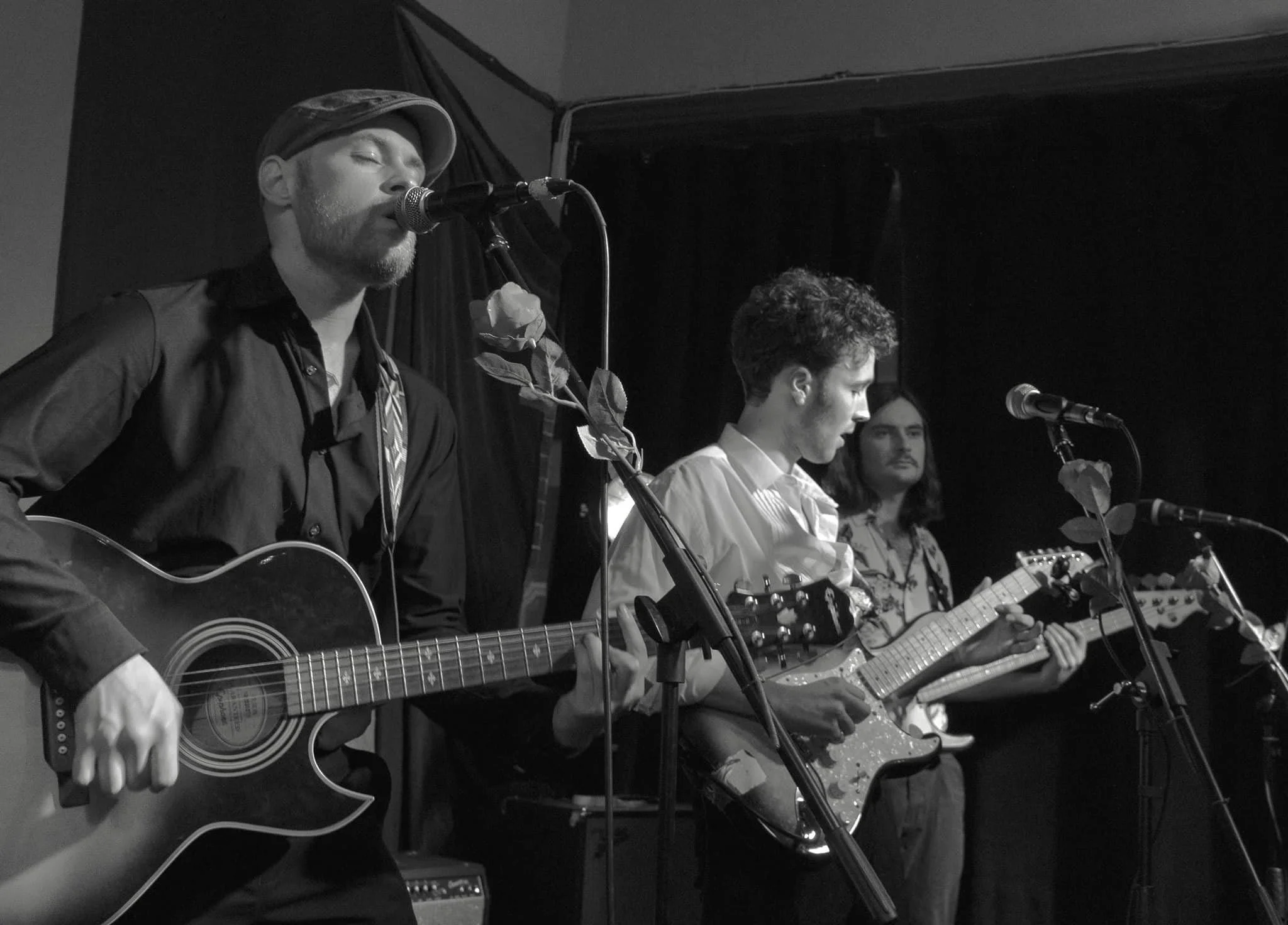 Black and white photo of four musicians performing on stage; the man on the left is singing into a microphone and playing an acoustic guitar, wearing a hat, and the three other men are playing electric guitars with serious expressions.