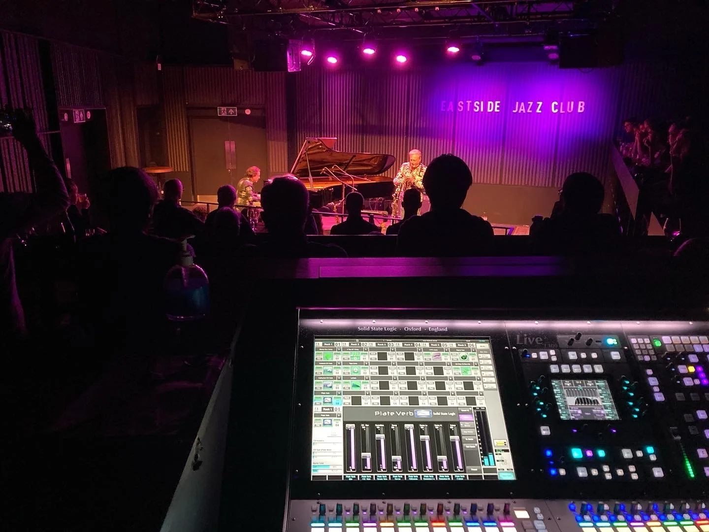View of a jazz performance at Eastside Jazz Club with two musicians playing the piano and saxophone on stage, purple lighting, audience seated watching, and a sound mixing console in the foreground.