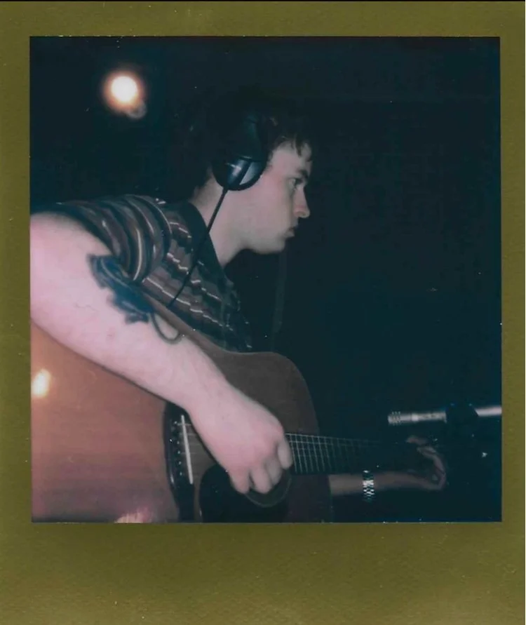 A young man with dark hair wearing headphones, playing an acoustic guitar, and looking off to the right in a dark room with a blurred light in the background.