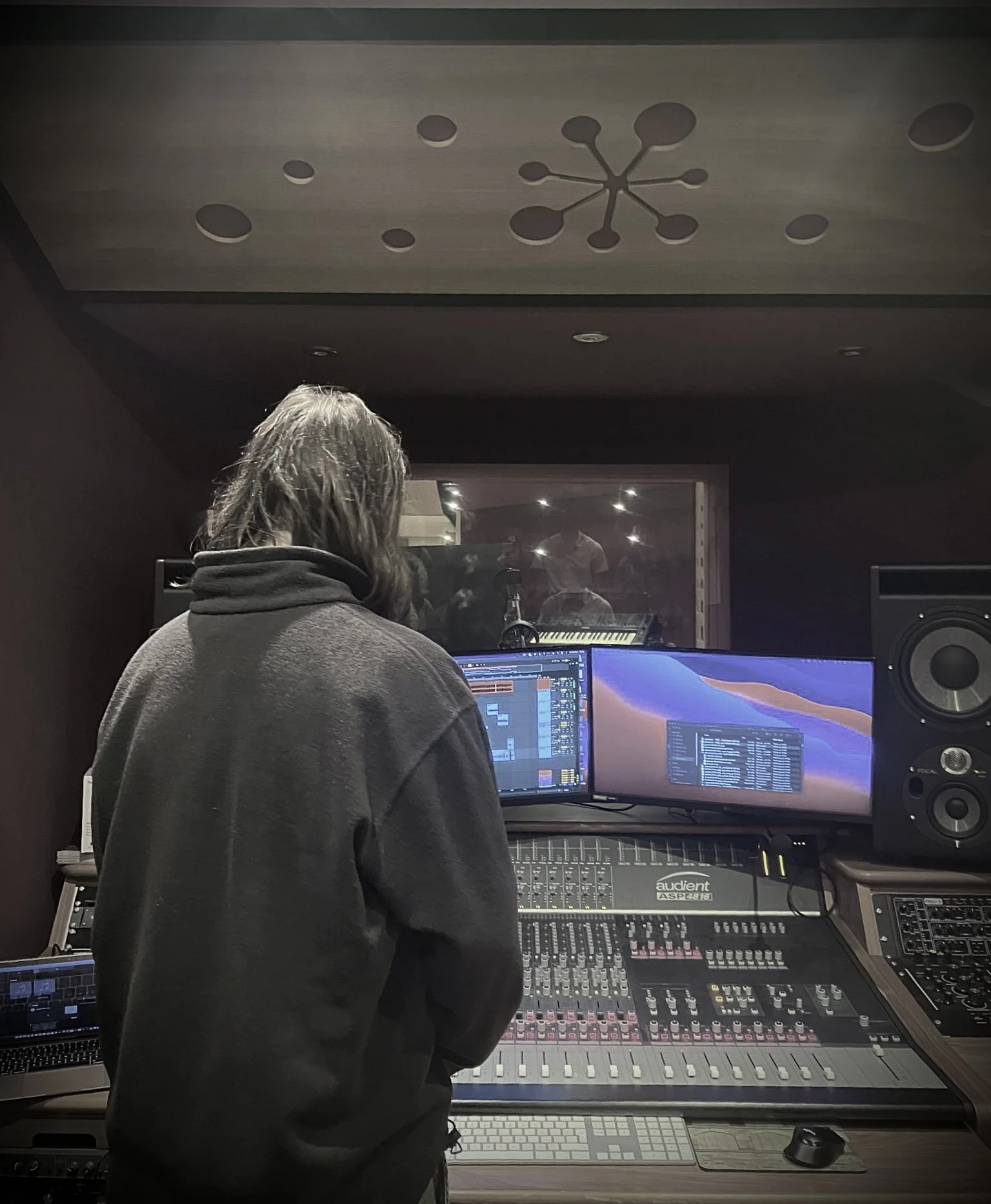 A person in a dark hoodie working in a recording studio with sound equipment, mixing console, multiple monitors, and a glass window showing another room with people.