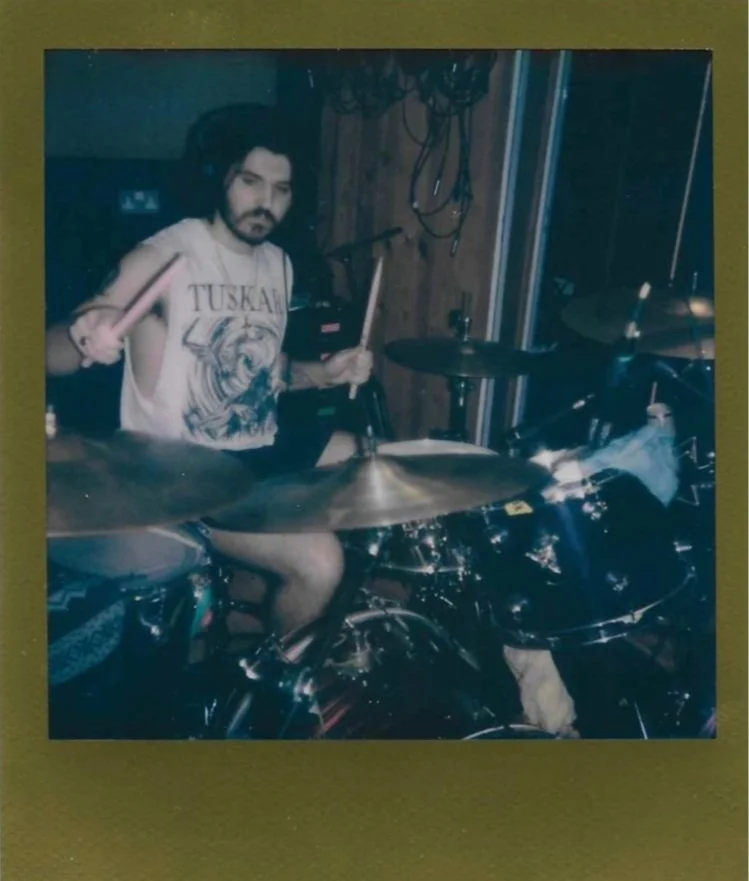 A person sitting behind a drum set holding drumsticks in a music studio or rehearsal space.