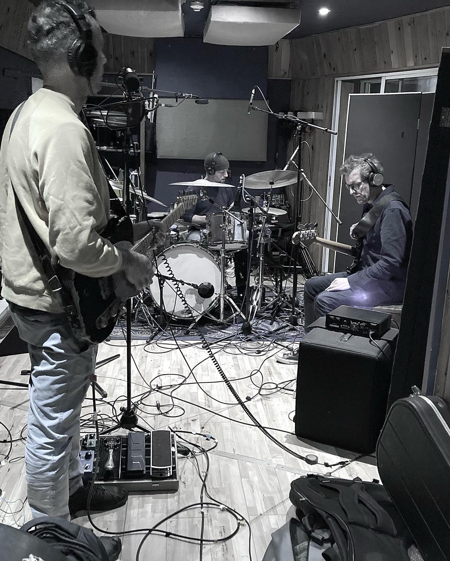 Three musicians in a recording studio. One man is standing while holding a guitar and wearing headphones. Two others are seated, playing drums and guitar, also with headphones. The room has wooden walls, a large door, and various recording equipment, wires, and amplifiers.