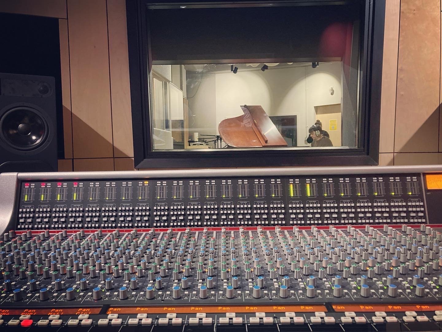 Sound mixing console in a recording studio with a view into the studio through a window, showing a grand piano and a person with headphones.