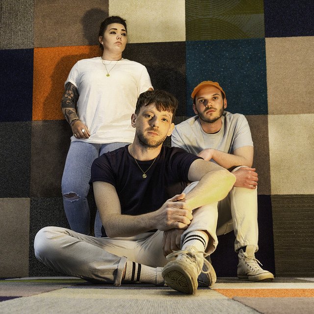 Three young adults posing indoors against a colorful, textured wall. One person with tattoos and dark hair is standing, another with a cap and beard is sitting, and a third with light hair is seated on the floor holding a shoe.