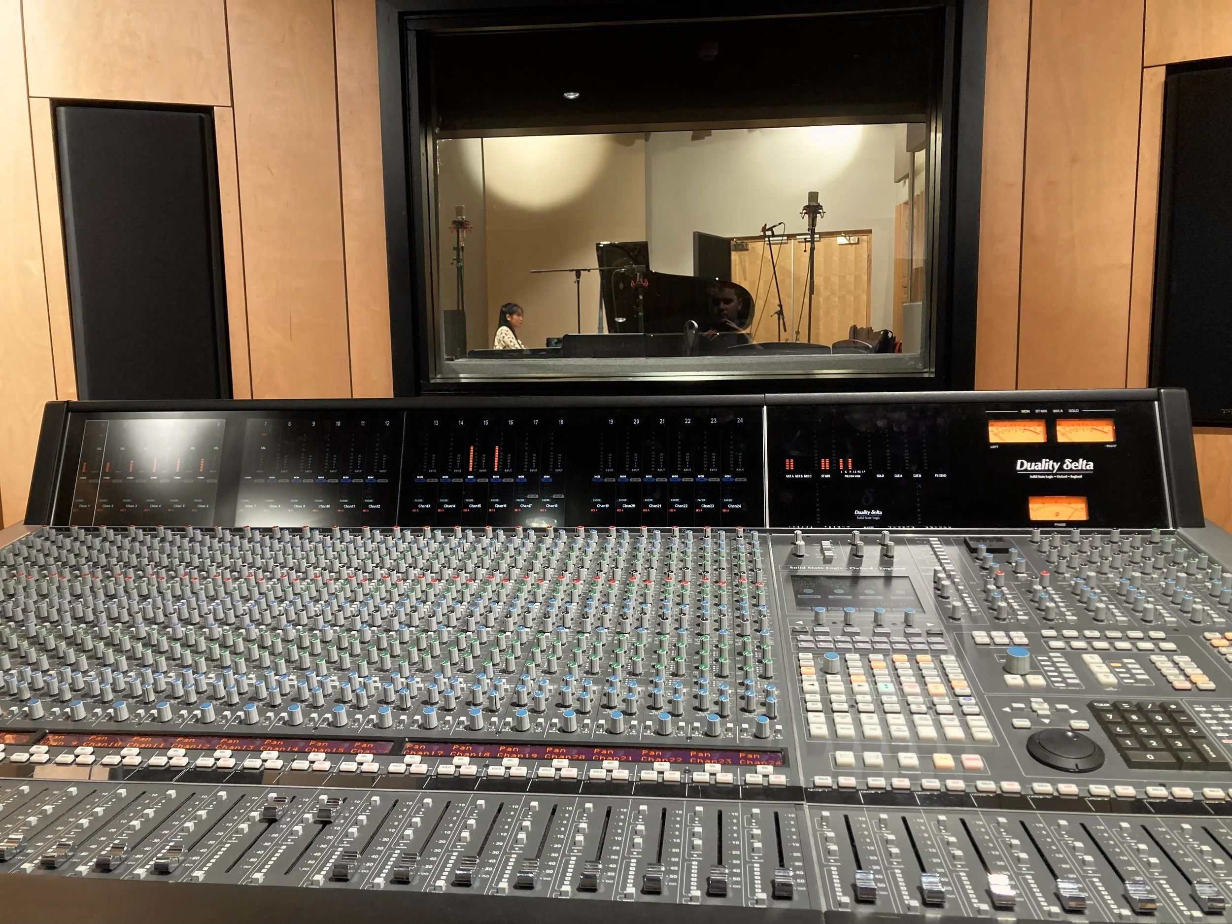 Sound mixing console with numerous controls and meters in a recording studio. Through a window, a woman is seen at a piano, in a room with wood-paneled walls.