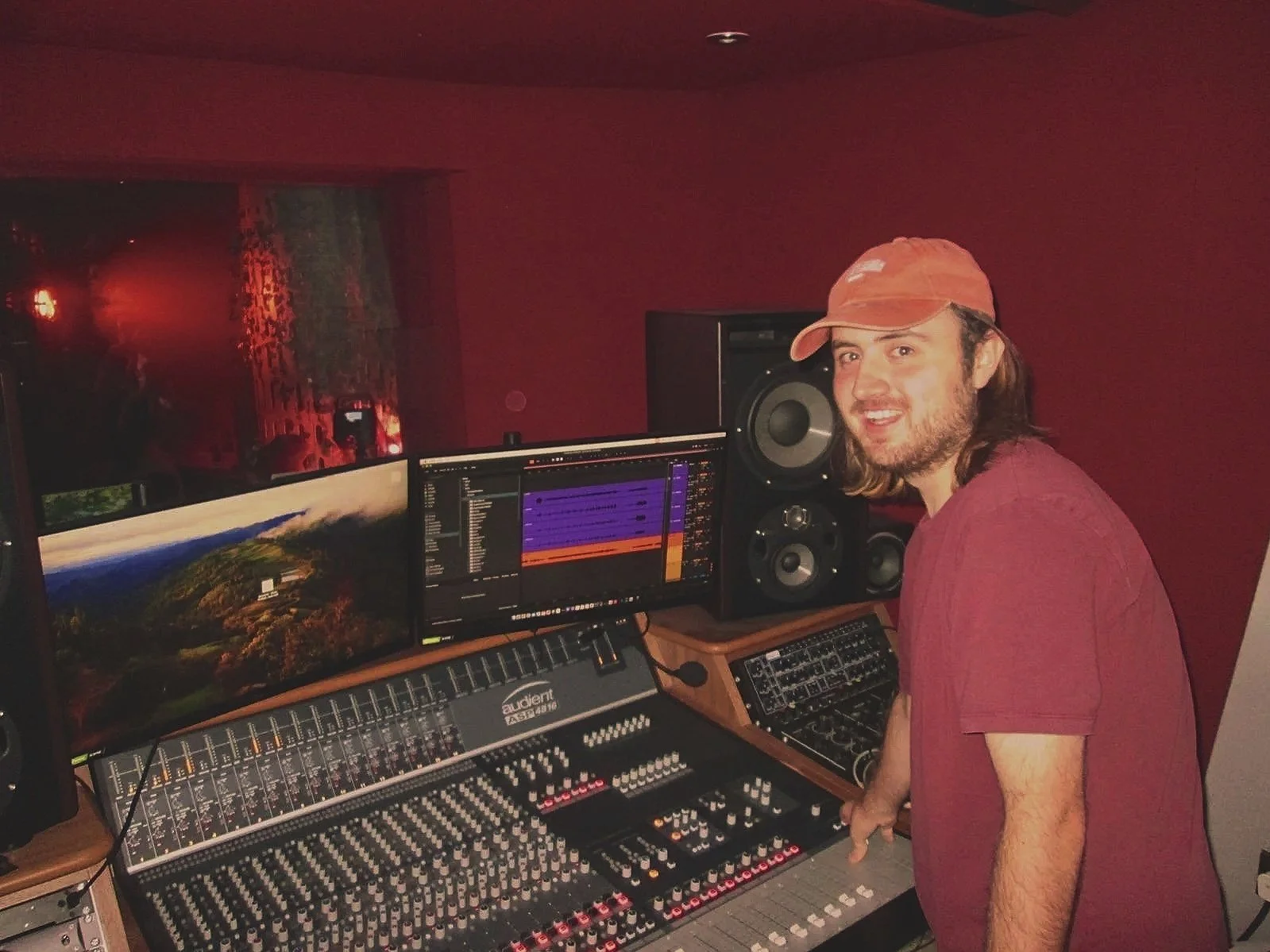 A man with shoulder-length hair and a beard wearing a red cap and red shirt in a music recording studio. He is standing in front of a mixing console, with large speakers and multiple monitors displaying audio editing software.
