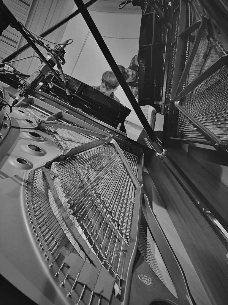 Two people are seen playing a grand piano from above, with the piano lid open, showing the strings and hammers inside.