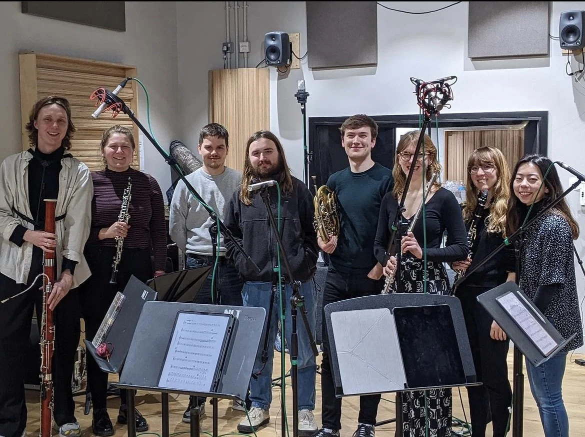 Group of nine young musicians in a recording studio, holding various musical instruments including a bassoon, oboe, French horn, and clarinet, standing behind music stands with sheet music.