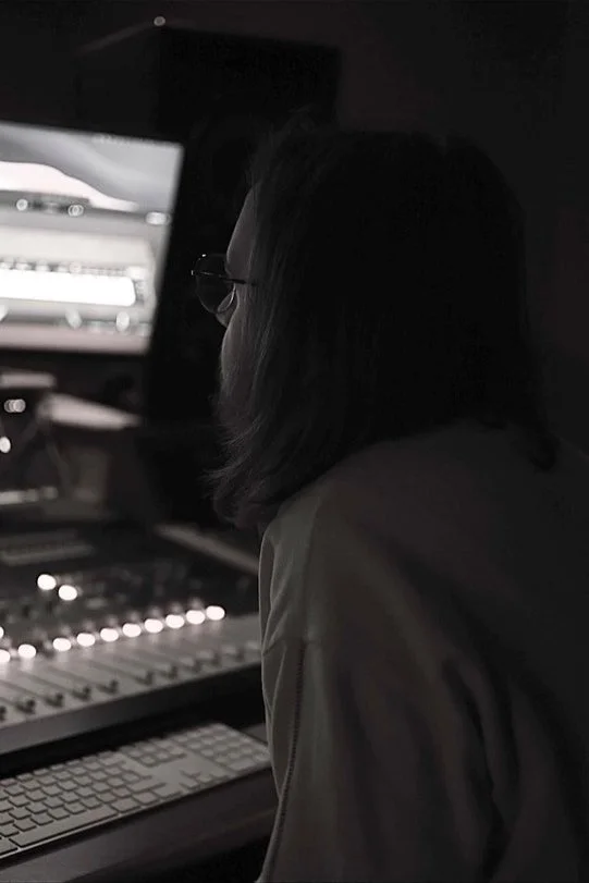 A woman with shoulder-length dark hair and glasses working on music or audio in a dark room with a computer monitor and audio equipment.