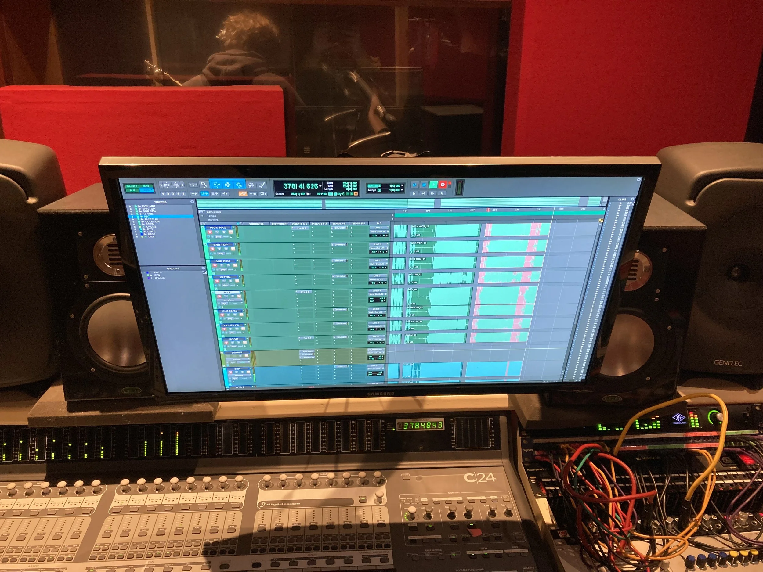 A music studio control room with a computer monitor displaying a digital audio workstation interface, a mixing console, and audio equipment with colorful cables.