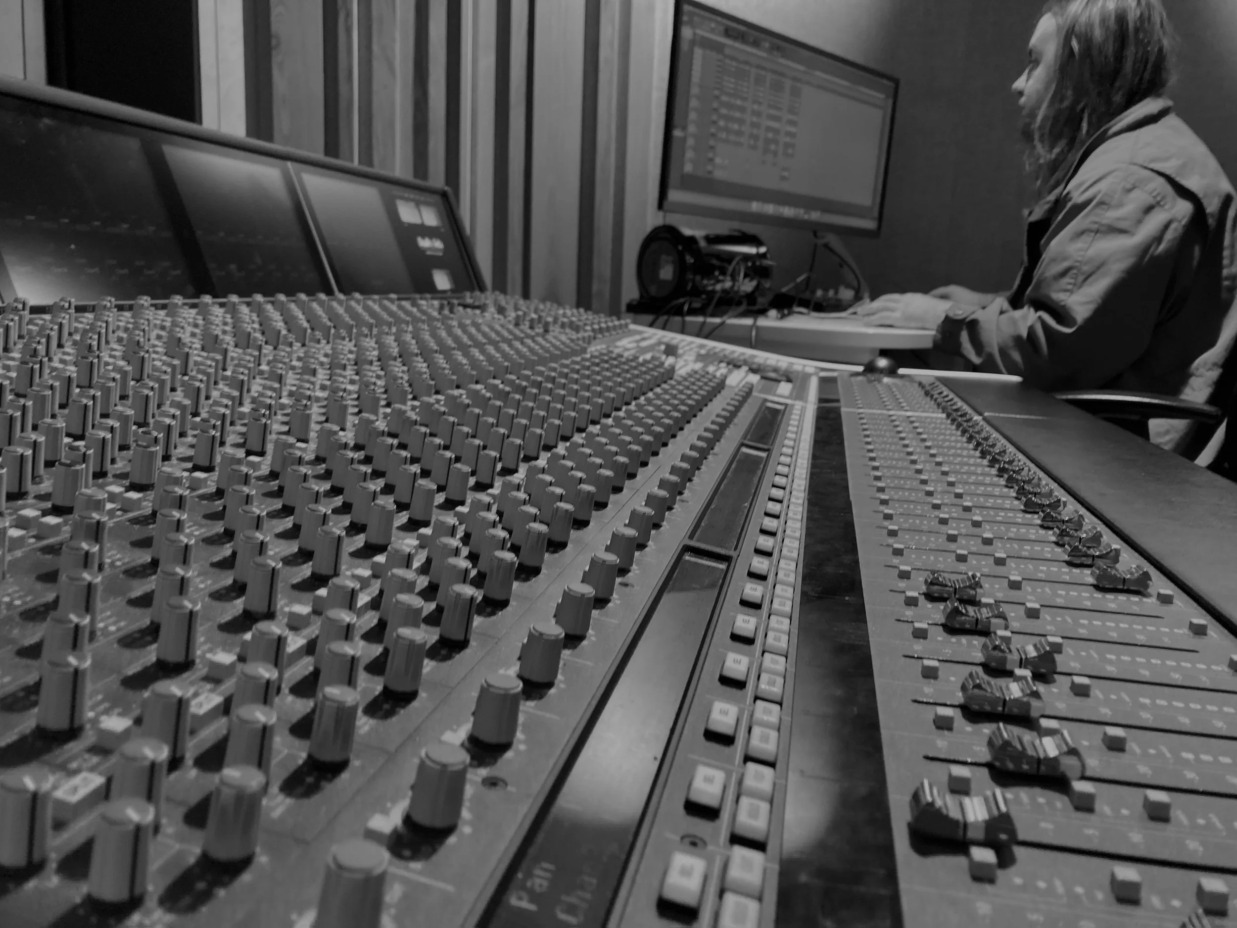 Black and white photo of a woman working with audio mixing equipment, including a large mixing console with numerous knobs and faders, and two computer monitors in a professional recording studio.
