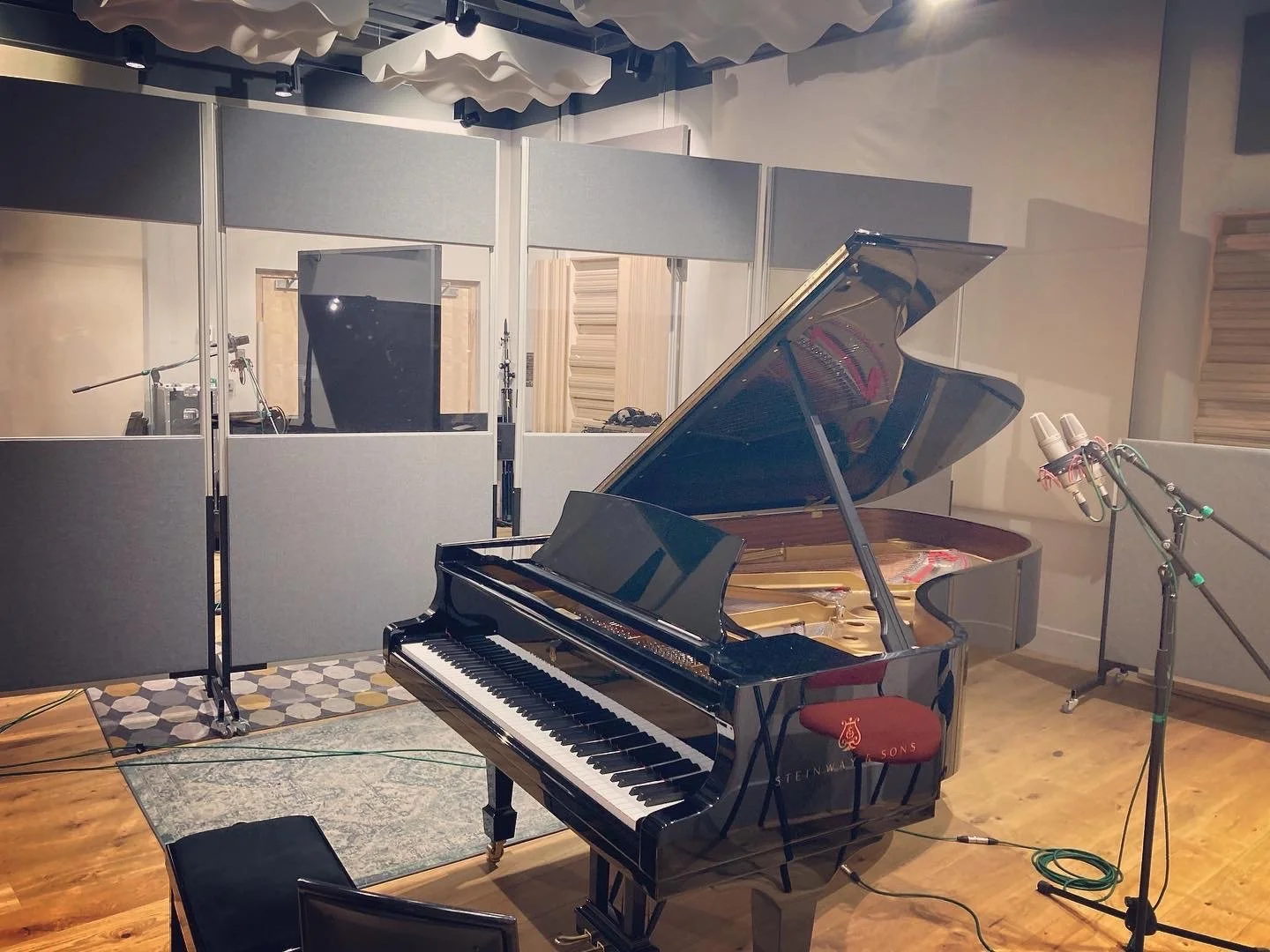 A black Steinway grand piano with an open lid in a recording studio, with microphones and audio equipment nearby, and soundproofed walls in the background.