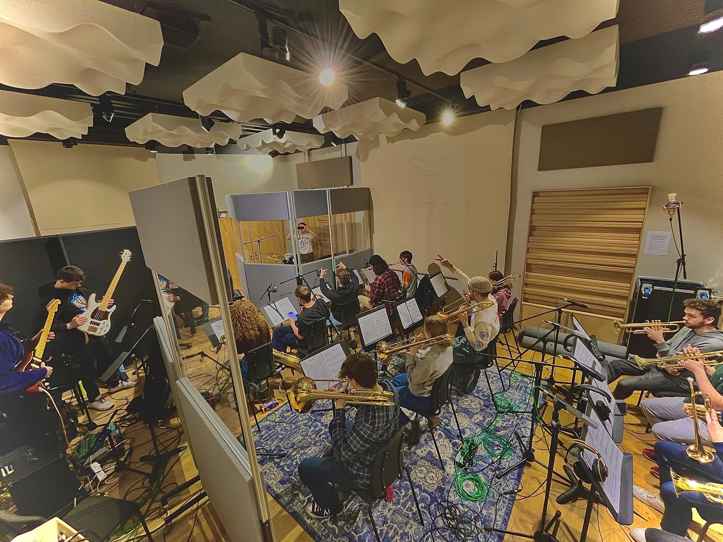 A music rehearsal or recording session in a studio with multiple musicians playing instruments like guitars, saxophones, and violins, and a producer or sound engineer wearing headphones overseeing the session.