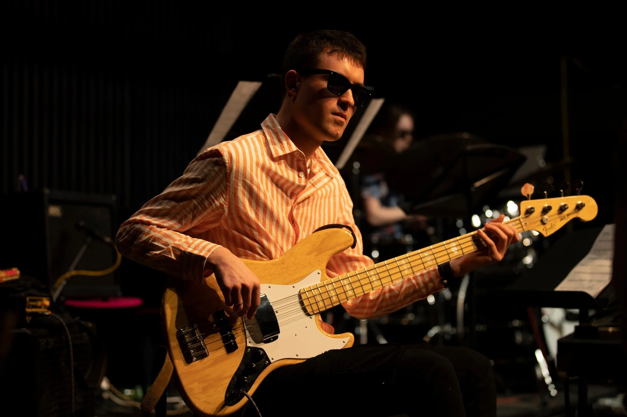 Man playing electric guitar in a dimly lit room with another musician visible in the background.