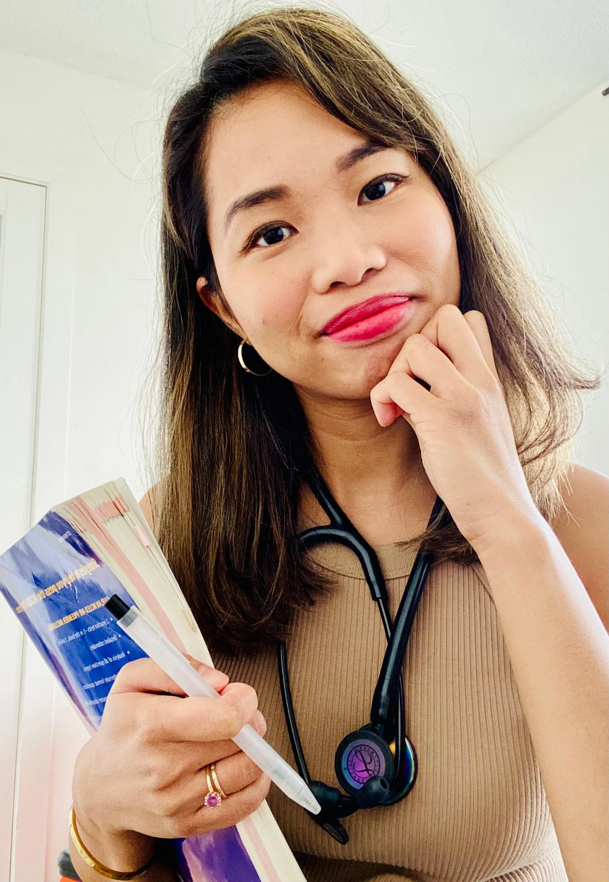 A young woman with brown hair and makeup, holding a book and pen, wearing a beige sleeveless top, gold hoop earrings, a gold ring with a pink stone, a gold bracelet, and a black stethoscope around her neck.