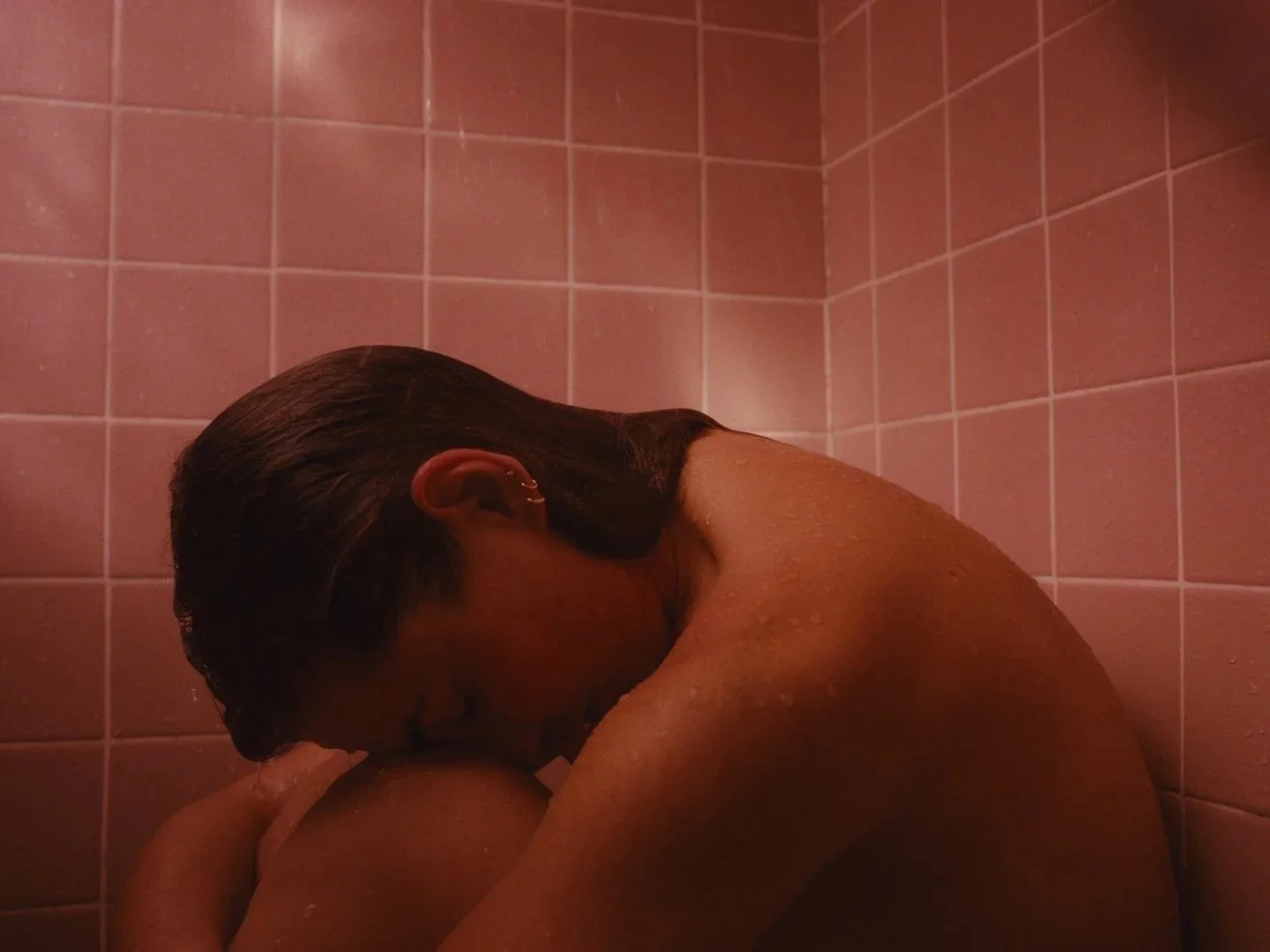 Person hugging their knees in a pink tiled shower area