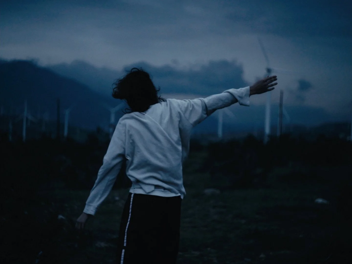 Person with dark hair wearing a white jacket, raising arm, in a dark outdoor landscape with wind turbines in the background during dusk or night.