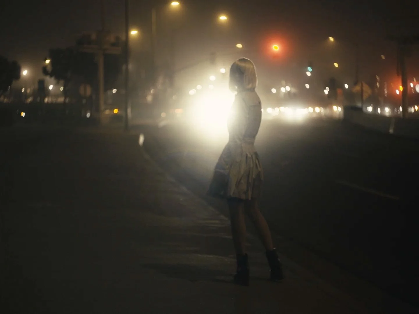A woman with blonde hair standing alone on a city street at night, facing away from the camera with bright headlights behind her, illuminated by streetlights and car lights.