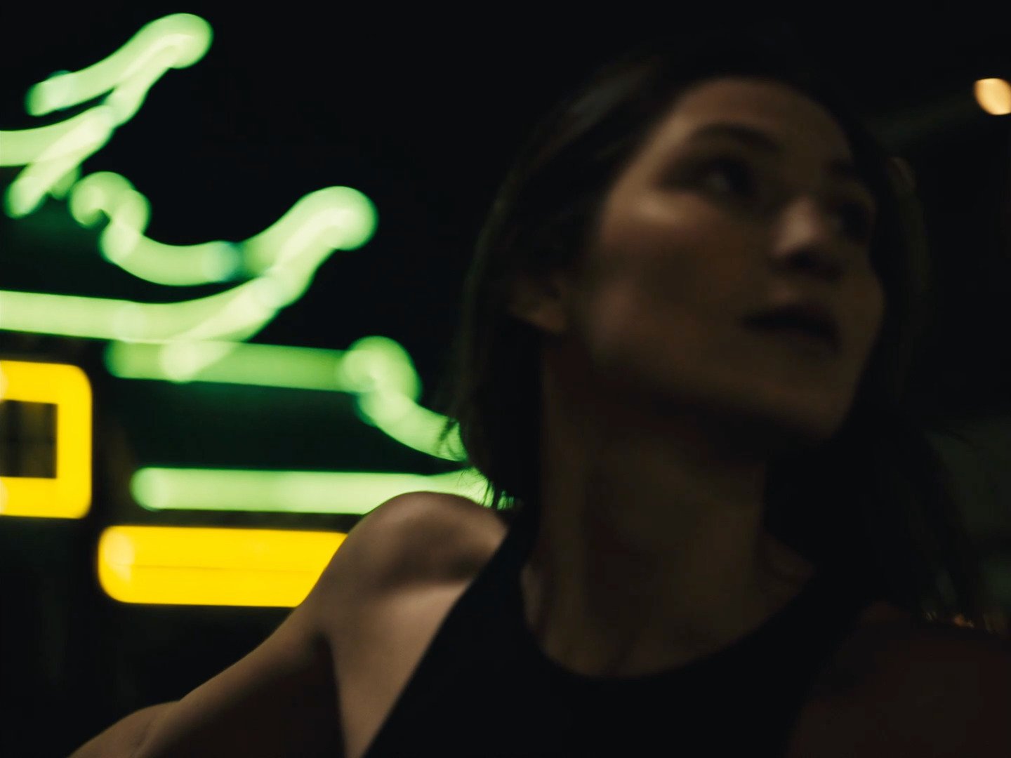 A woman with dark hair is partially visible in a dark environment, with neon lights in green and yellow in the background.