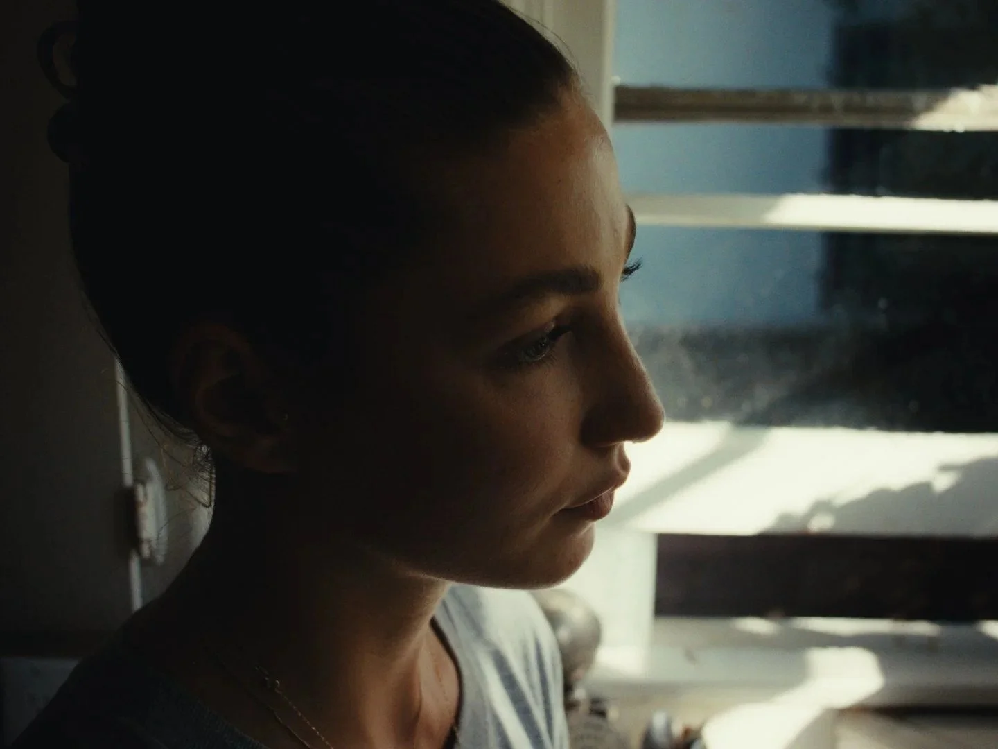 Side profile of a woman with dark hair pulled back, looking down, near a window with sunlight streaming through the blinds.