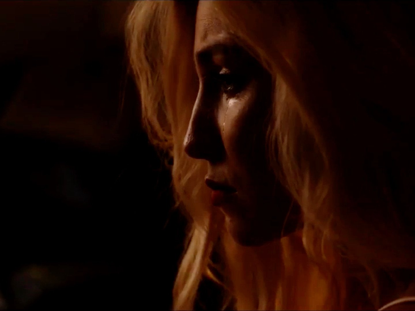 A close-up side profile of a woman with long, wavy blonde hair, with dim lighting creating shadows on her face.