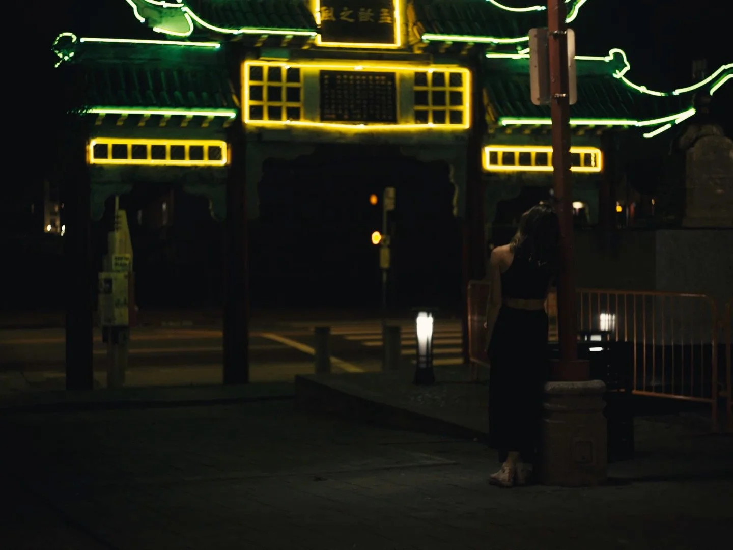 A woman standing under streetlights near a traditional Chinese gate illuminated with green and yellow neon lights at night.