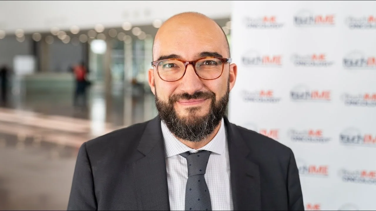 A man with a beard and glasses wearing a black suit and tie, smiling, at an event with a blurred background and a step-and-repeat banner that says 'TECHMARE'.