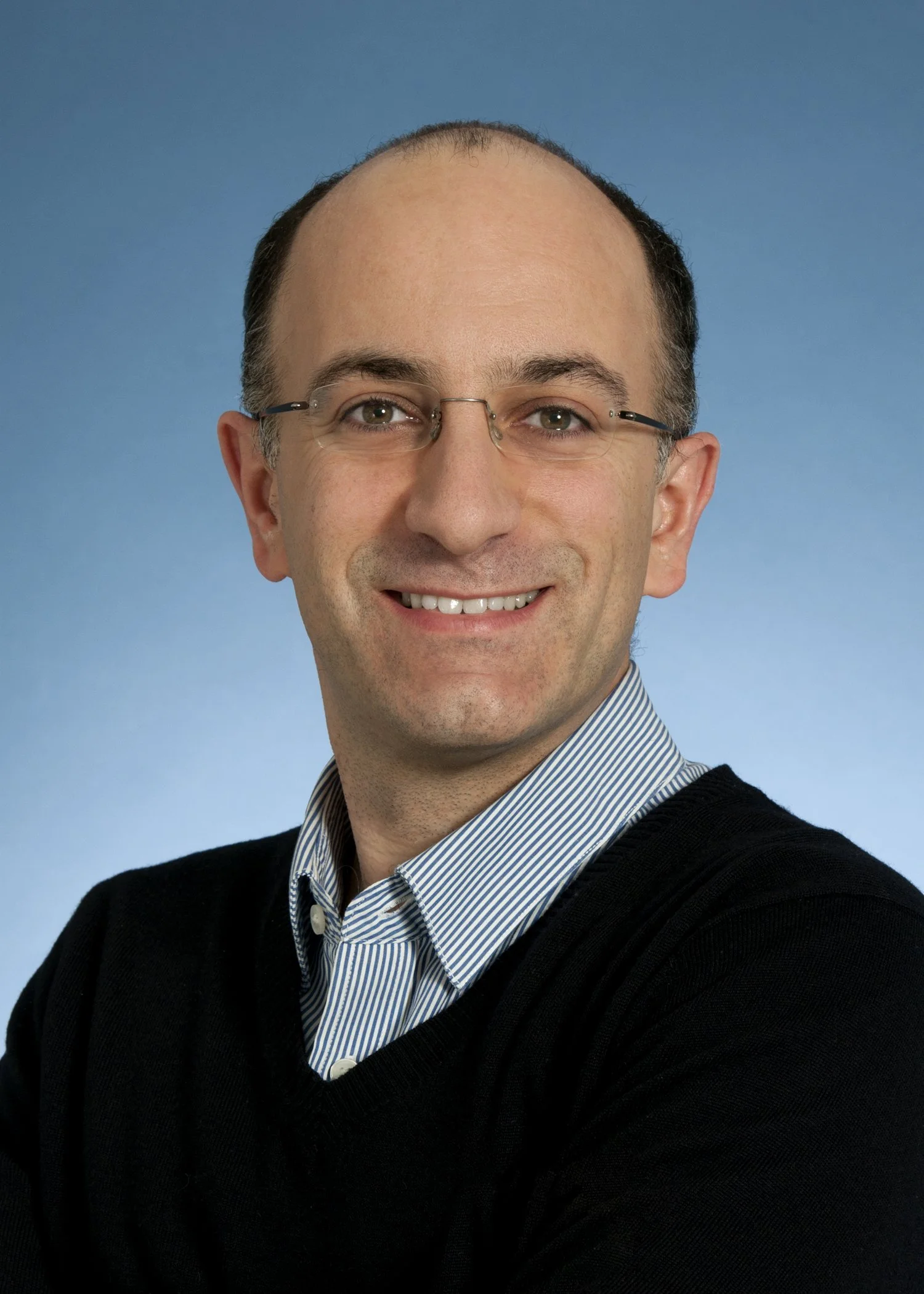 Portrait of a smiling man with glasses, wearing a striped shirt and black sweater, against a blue background.