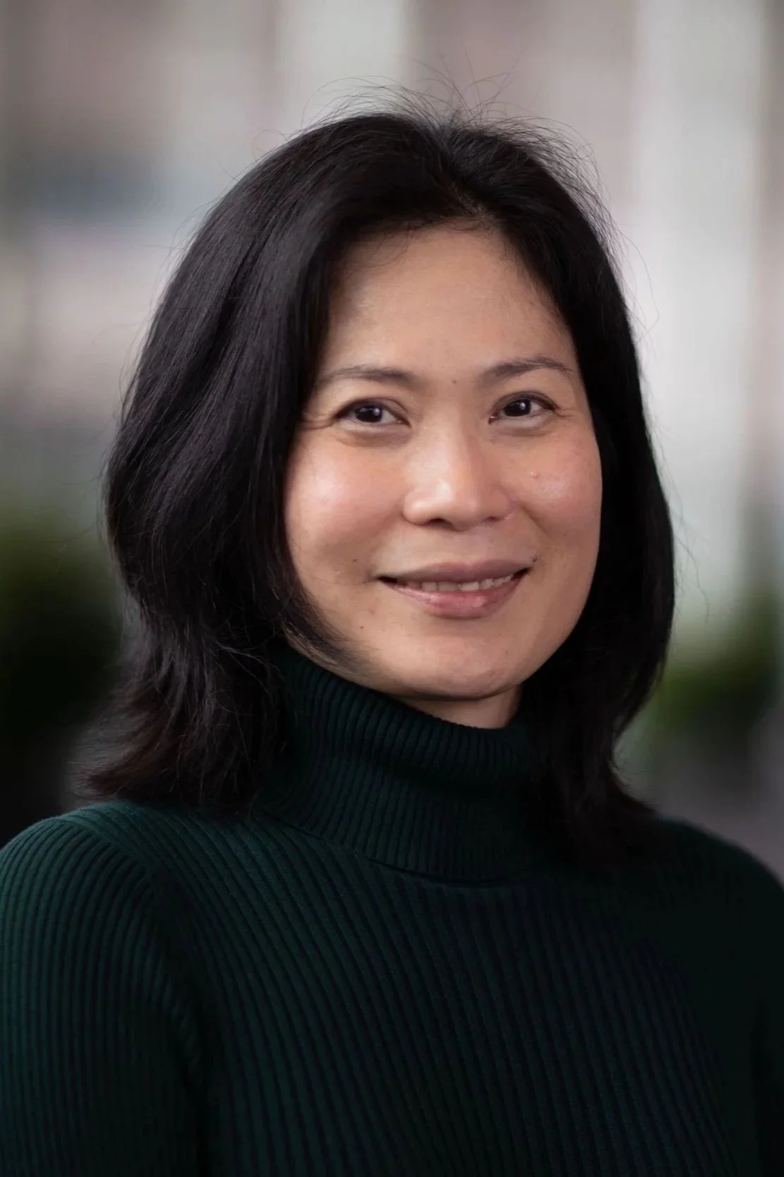A woman with black hair wearing a dark green ribbed turtleneck sweater, smiling, with a blurred indoor background.