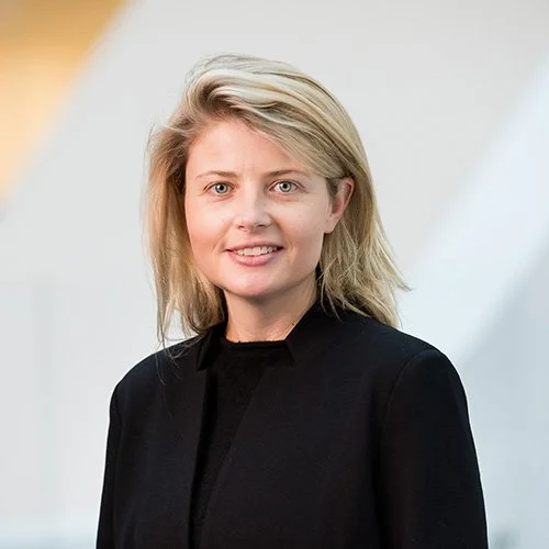 A portrait of a woman with blonde hair, wearing a black blazer, smiling at the camera, with a blurred indoor background.