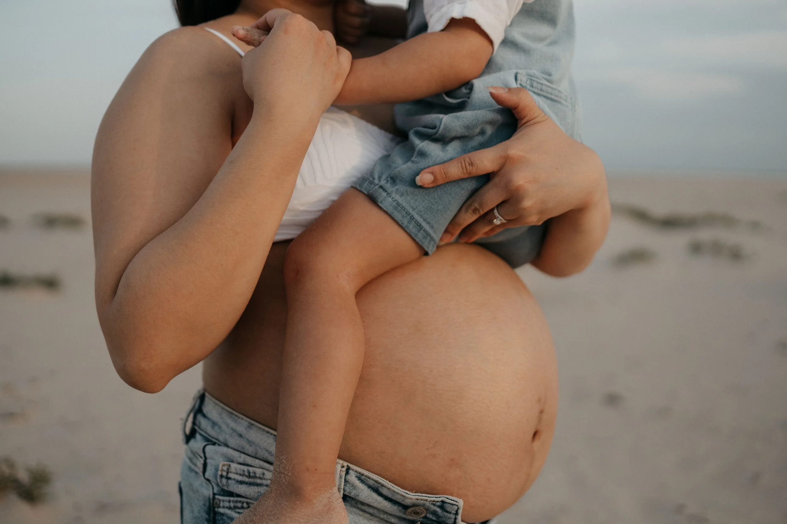 A pregnant woman holding a small child on a beach. The woman is wearing denim shorts and a white top, and the child is wearing a white shirt and denim shorts. The woman is cradling the child with one arm and holding the child's hand with the other.