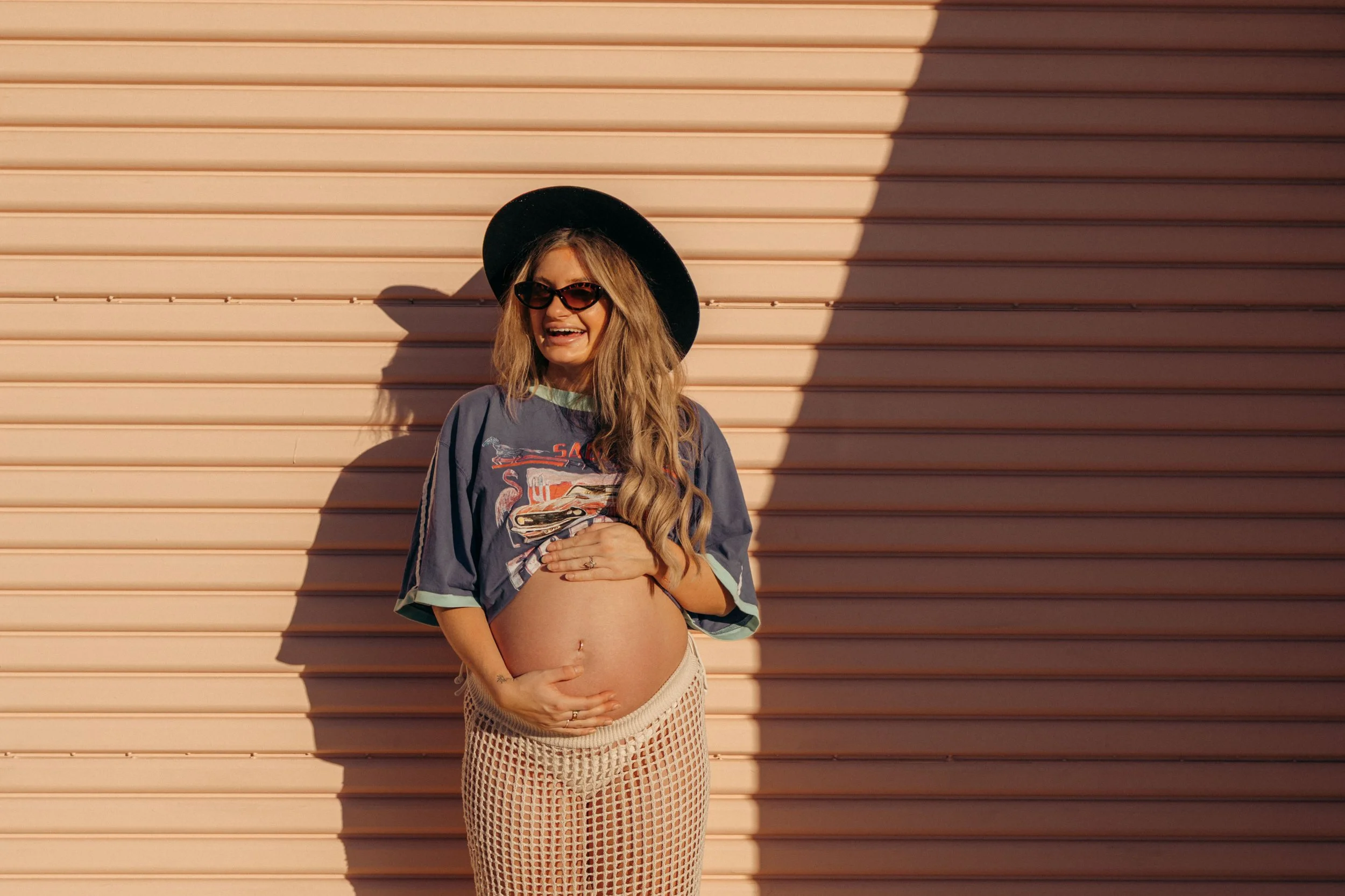 A pregnant woman standing in front of a pinkish garage door, wearing sunglasses, a black wide-brim hat, a vintage graphic t-shirt, beige fishnet pants, and smiling.