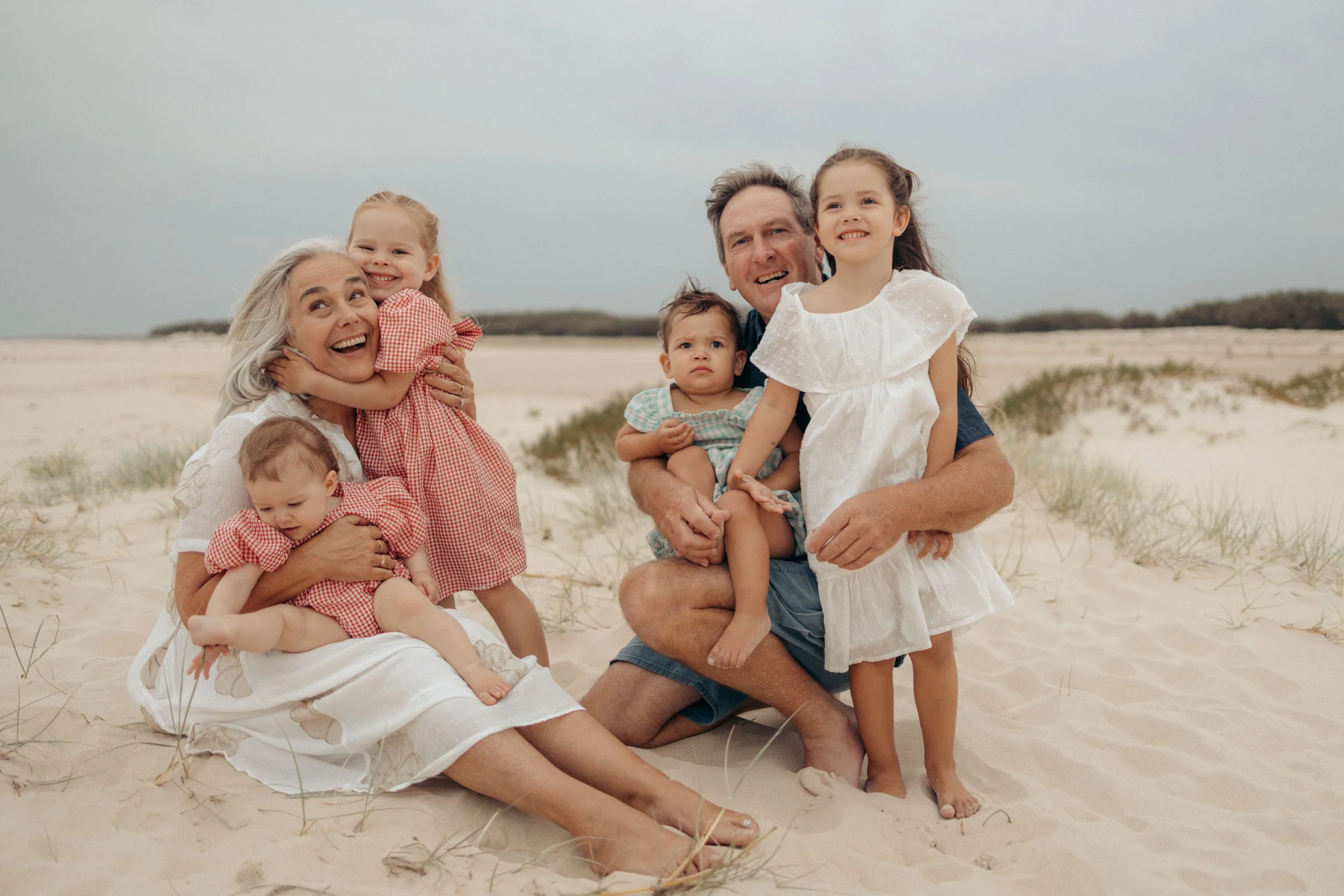 Family of six sitting on sandy beach with dune grasses, smiling and enjoying a cloudy day.