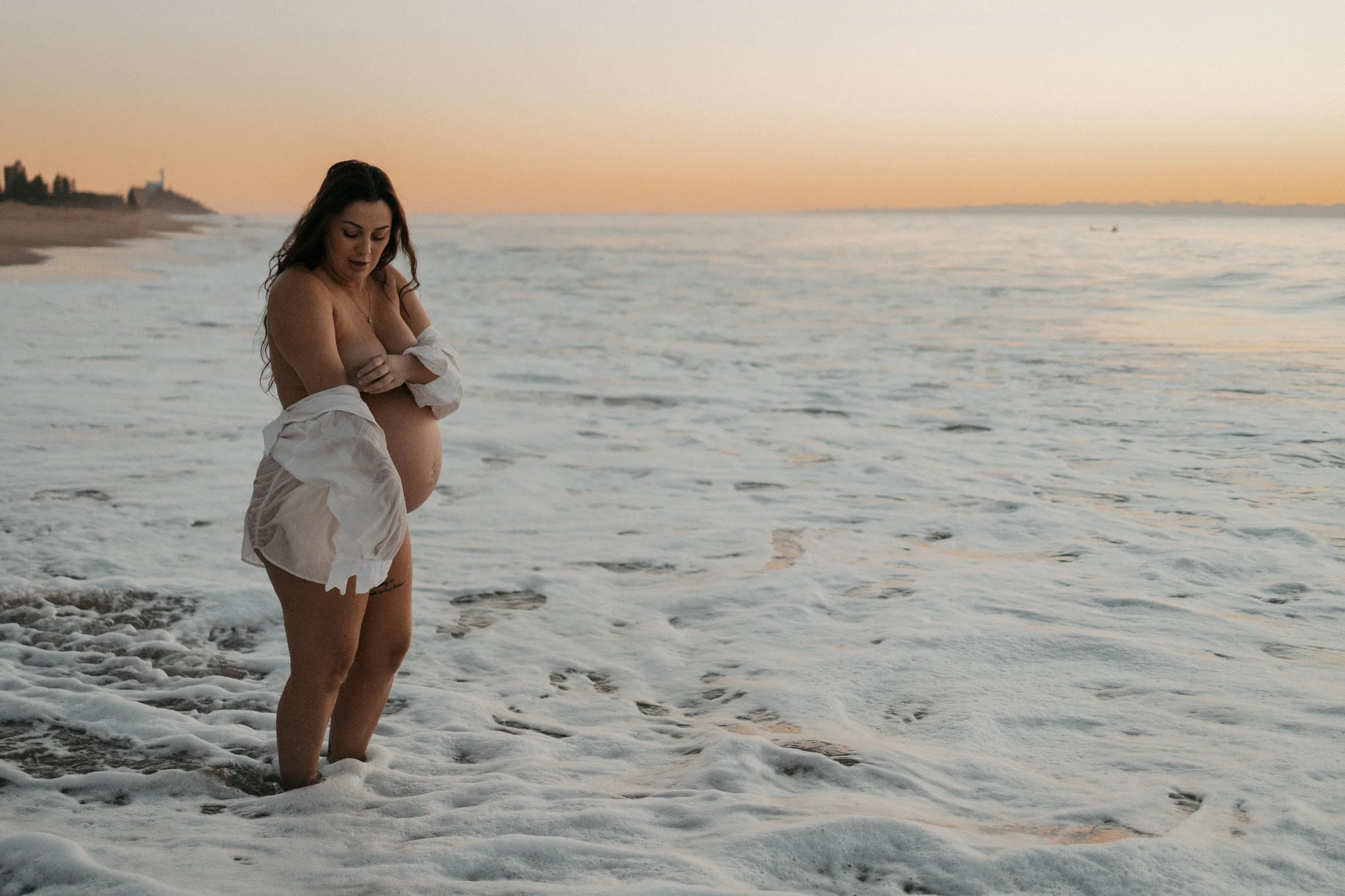 Pregnant woman standing in the ocean at sunset, wearing a white shirt draped off her shoulders.