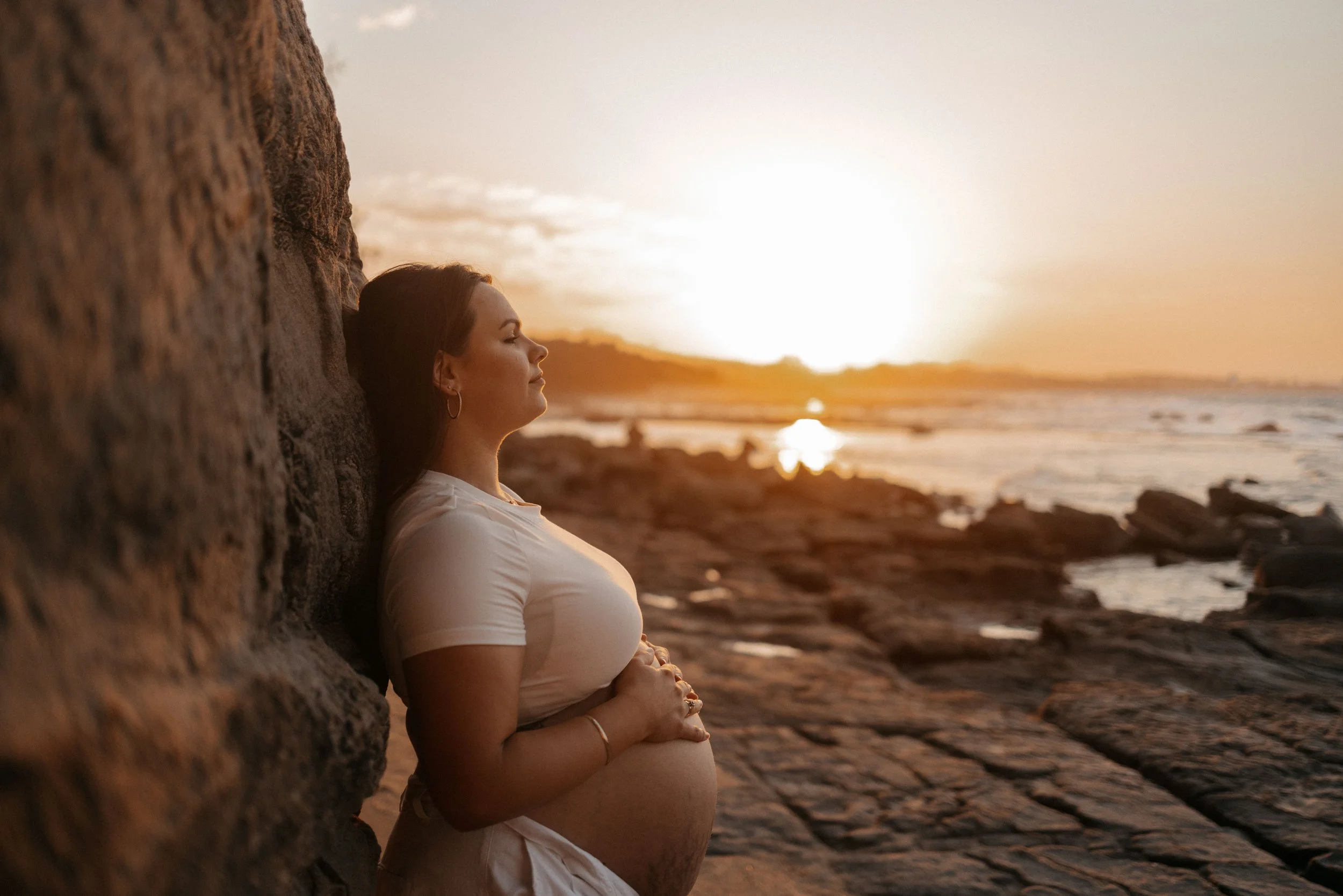 A pregnant woman leaning against a large rock on a beach at sunset, with her eyes closed and hands on her belly.
