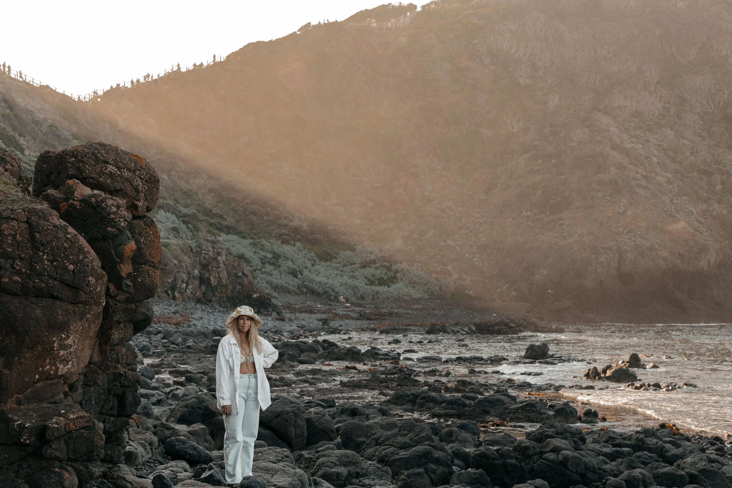 A woman in white pants, a white jacket, and a wide-brimmed hat standing on a rocky beach with large rocks and ocean waves, with hills and sunlight in the background.