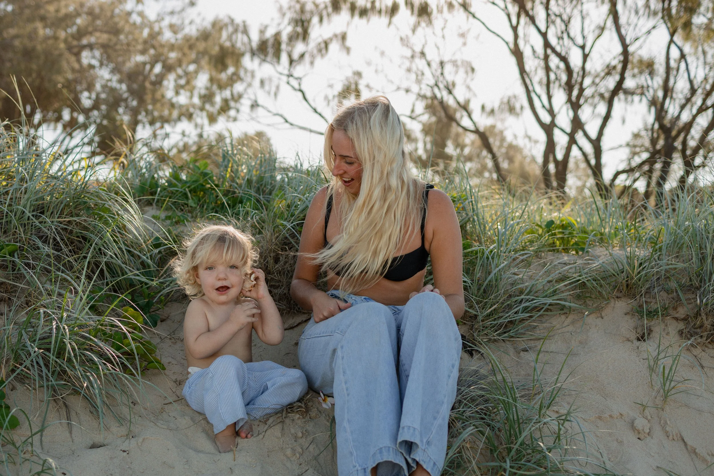 A woman holding a young girl on a sandy beach during sunset, with footprints in the sand and trees in the background.