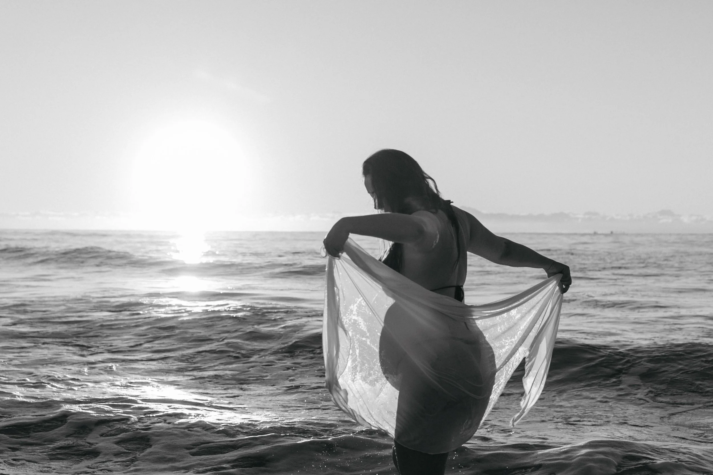 A woman standing in the ocean at sunset, holding a semi-transparent piece of fabric, with the sun setting on the horizon.