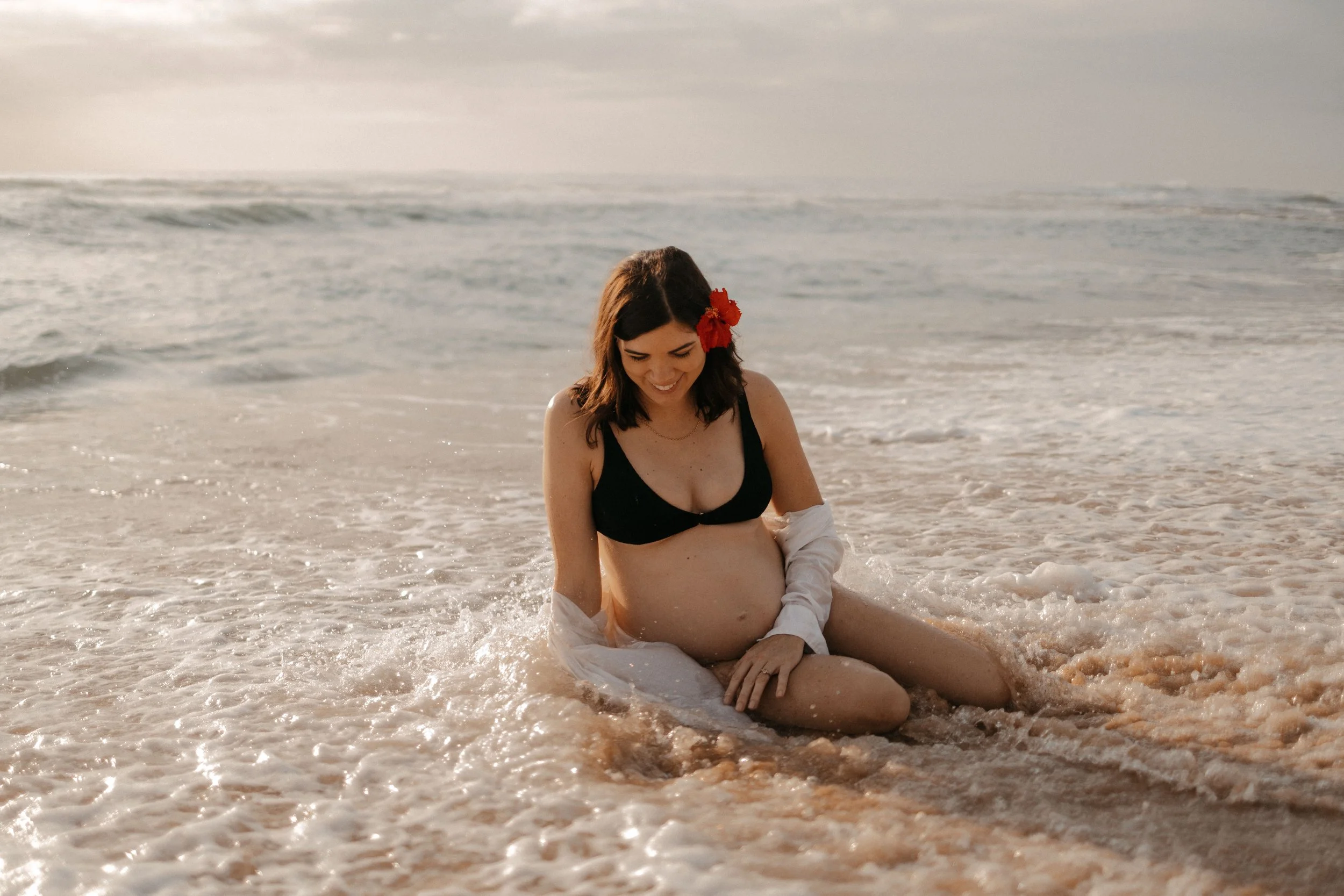 A pregnant woman with a red flower in her hair sitting in the shallow surf at the beach, smiling down.