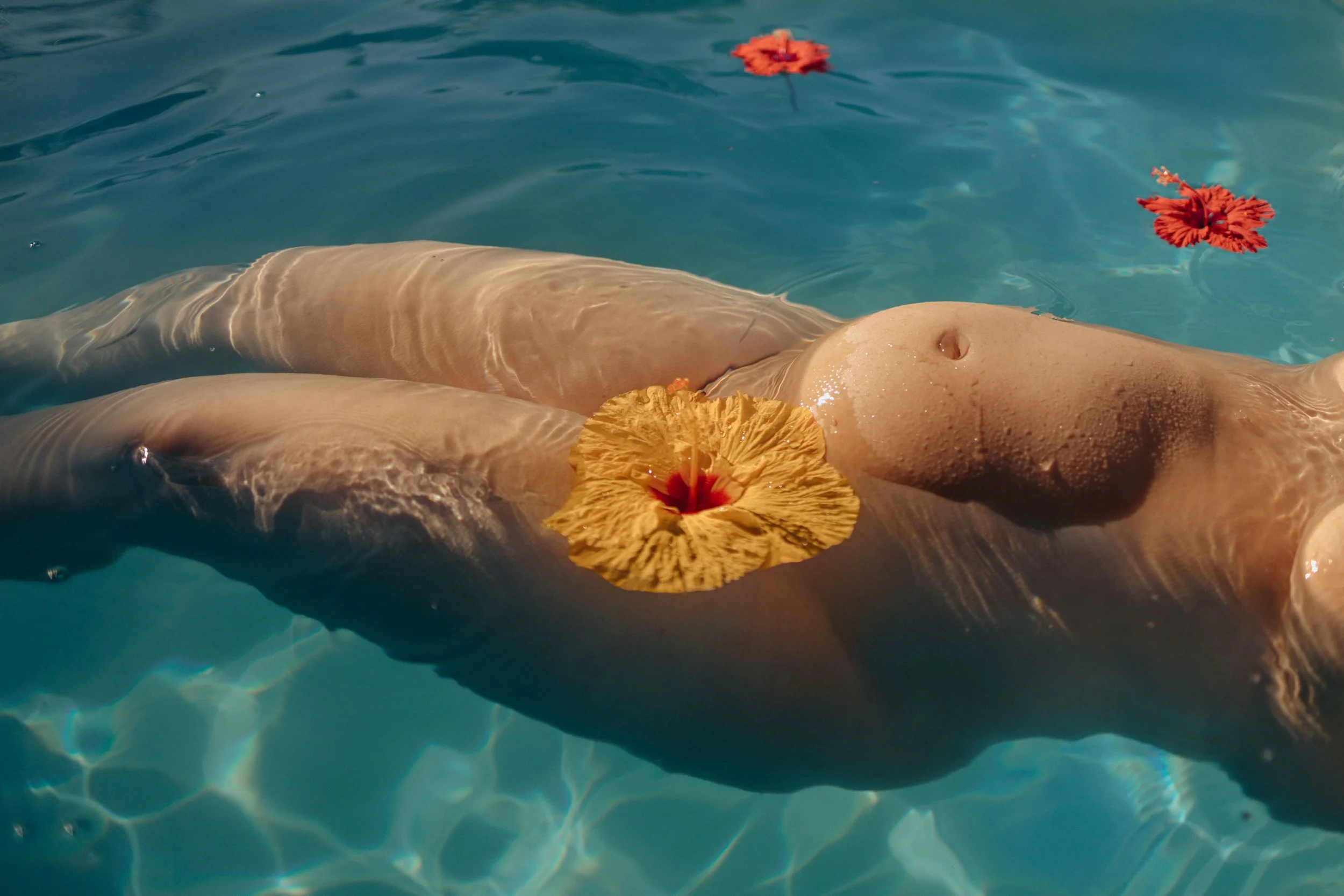 A person floating in a pool with three colorful hibiscus flowers on their body and in the water.