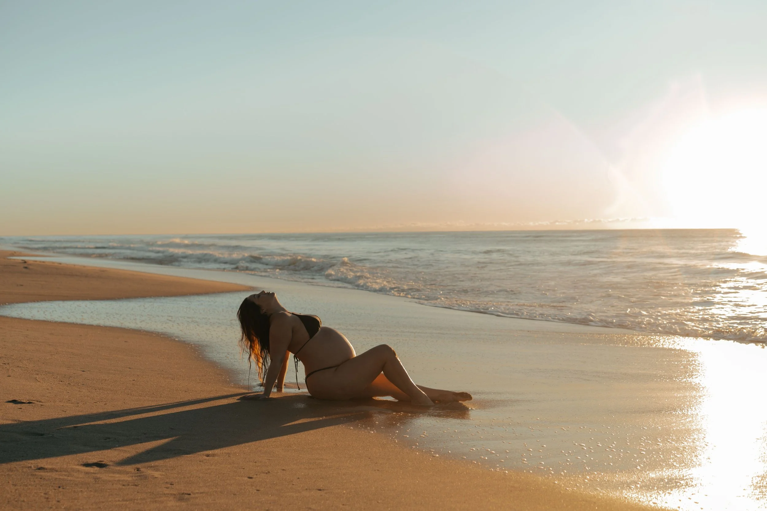 A woman in a black bikini reclining on a sandy beach near the water during sunset.